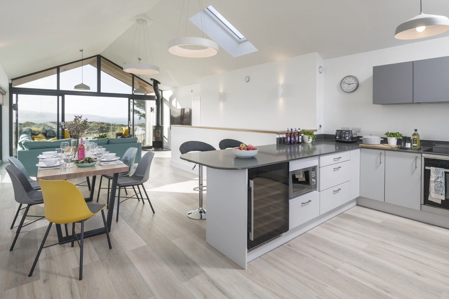 Open plan kitchen, dining room and lounge. White and grey kitchen fixtures. Wooden dining table with chairs. Teal sofa.
