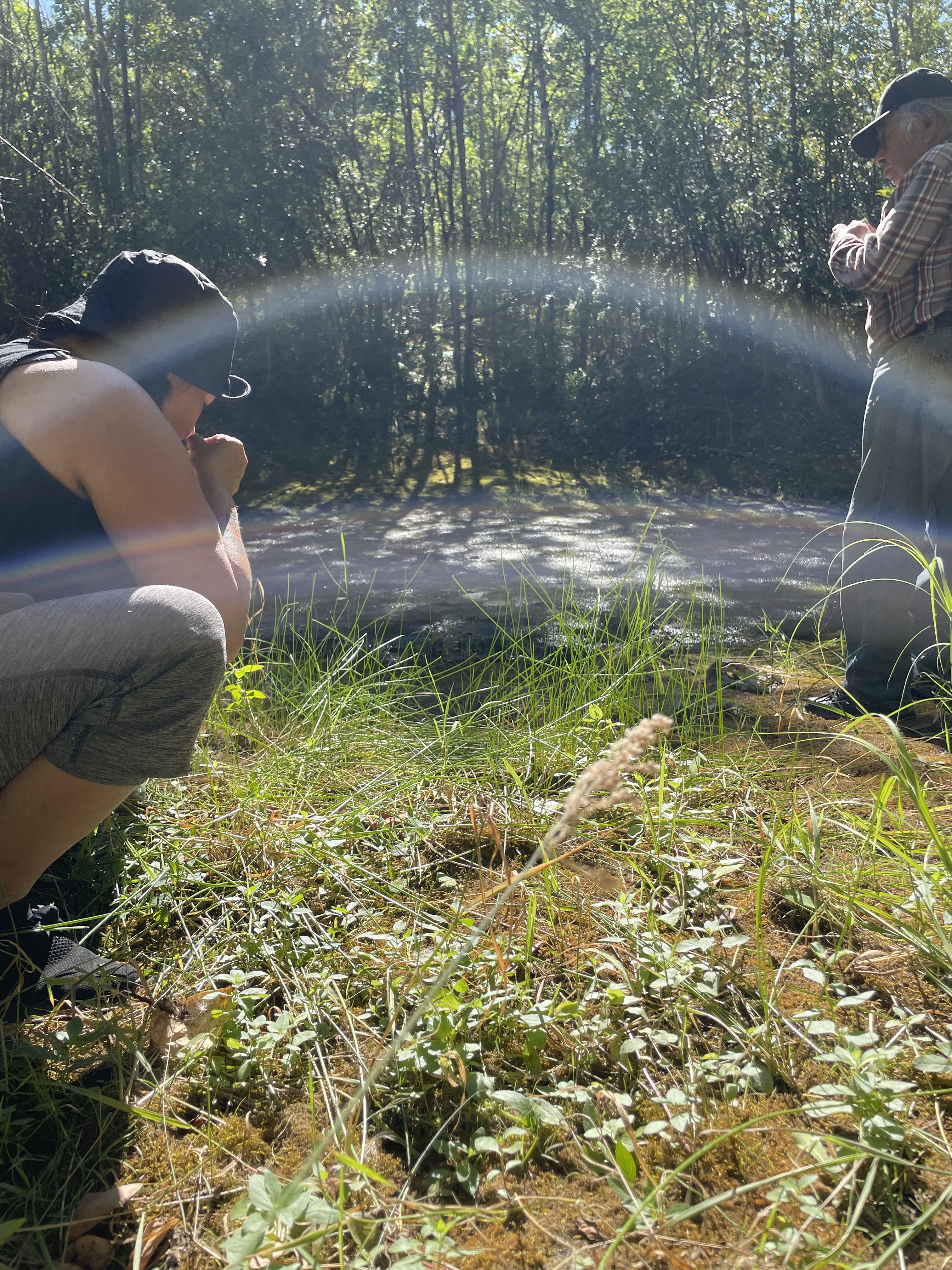 A sunny photo of Kiona crouching in green grass beside a forest path. There are trees in the background and Kiona's Moshom stands beside Kiona to the right.