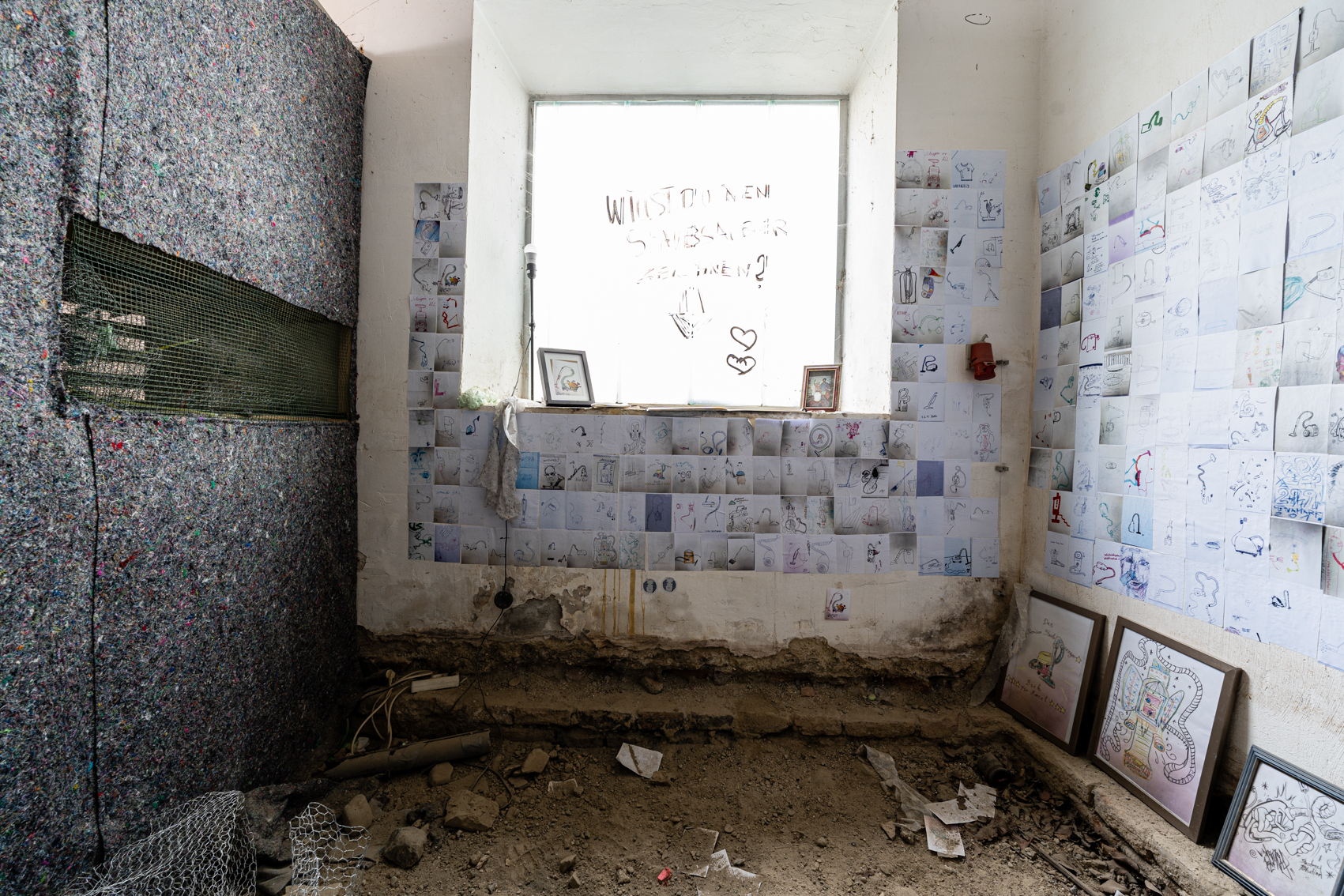 Bild 2: Im inneren des Raumes befindet sich ein gro&szlig;es quadratisches Glasfenster auf einer Anh&ouml;he. Darauf steht: Willst du einen Staubsauber malen? An der rechten Wand und rund um das Fenster, h&auml;ngen viele kleine Zeichnungen von Staubsaugern.