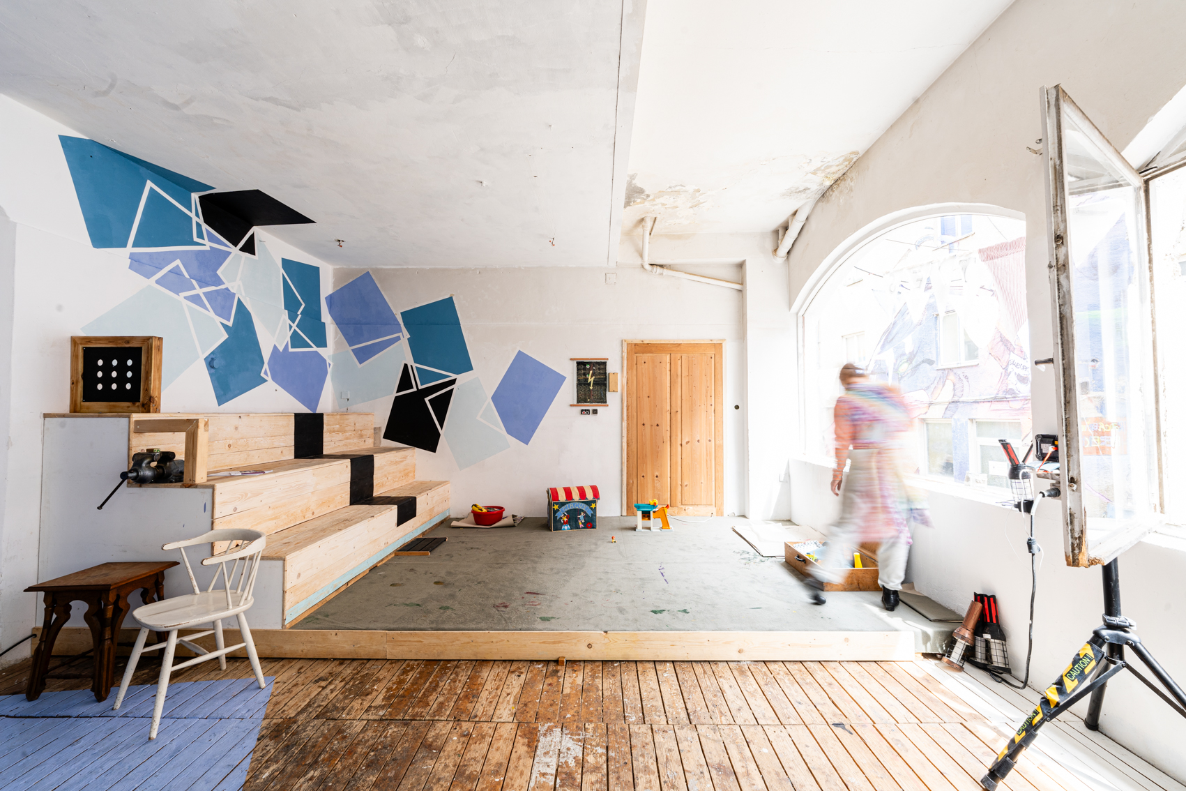 Bild 4 Cafe Eder: Das Bild zeigt den leicht erh&ouml;hten Spiel- und B&uuml;henenbereich des Cafes. Links der B&uuml;hne gibt es eine dreistufige Sitzm&ouml;glichkeit aus Holz. Der Rest der B&uuml;hne ist mit Teppichboden verkleidet. An den W&auml;nden links sind Rechtecke in verschiedenen Blau- und  Lila-T&ouml;nen an die Wand gemalt.