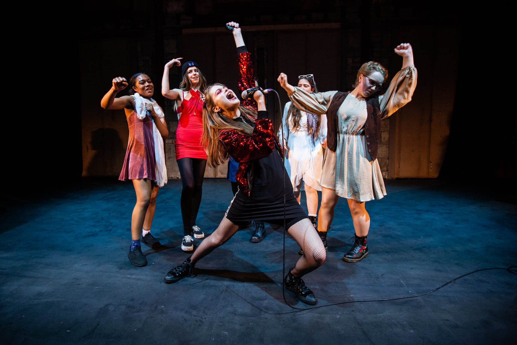 A group of young women/femmes dance onstage. In front of them, another yells triumphantly into a microphone, her arm in the air.