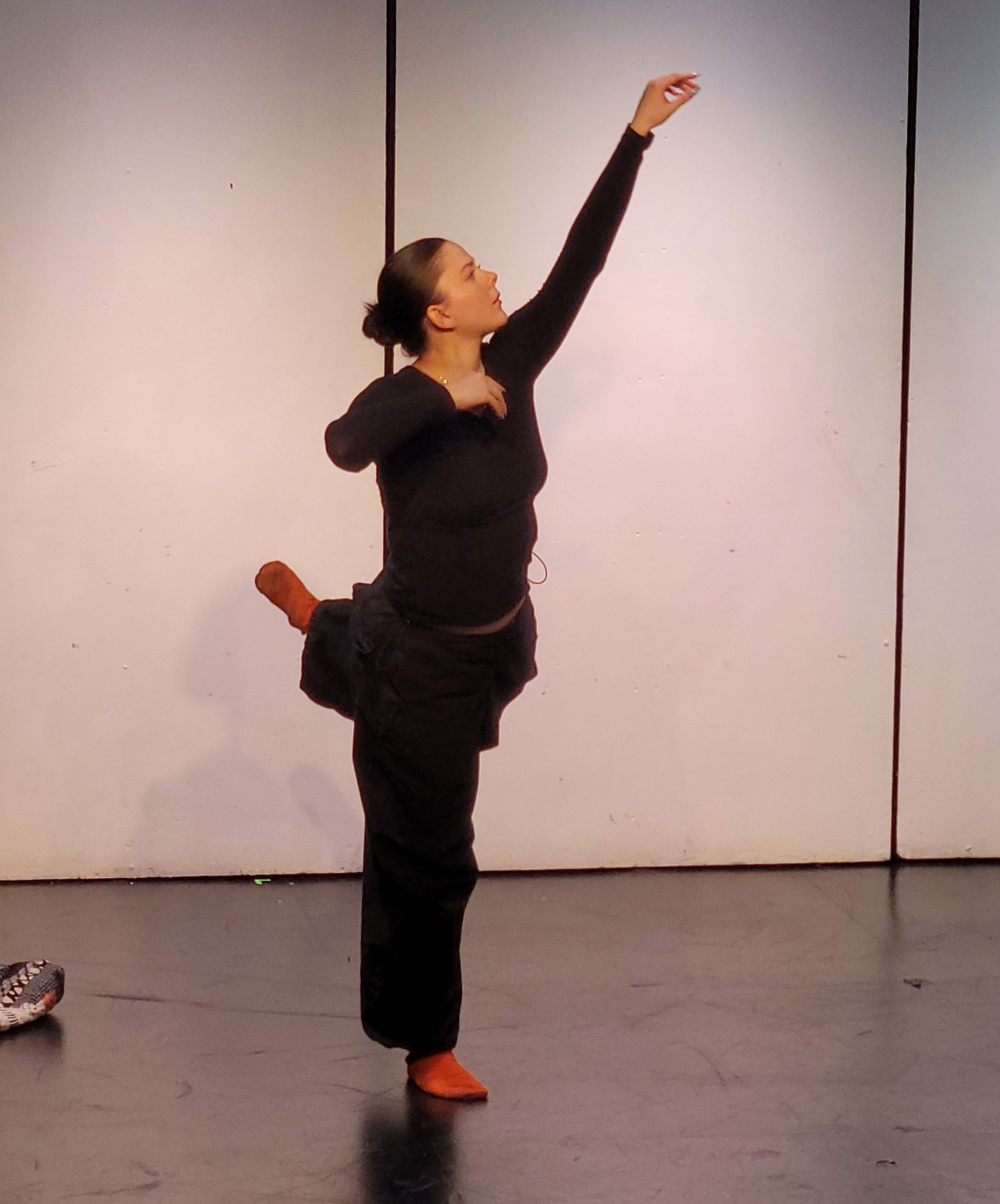 Rachel balances on one leg, reaching an arm up overhead as if plucking something out of the air. She wears all black with bright orange socks.