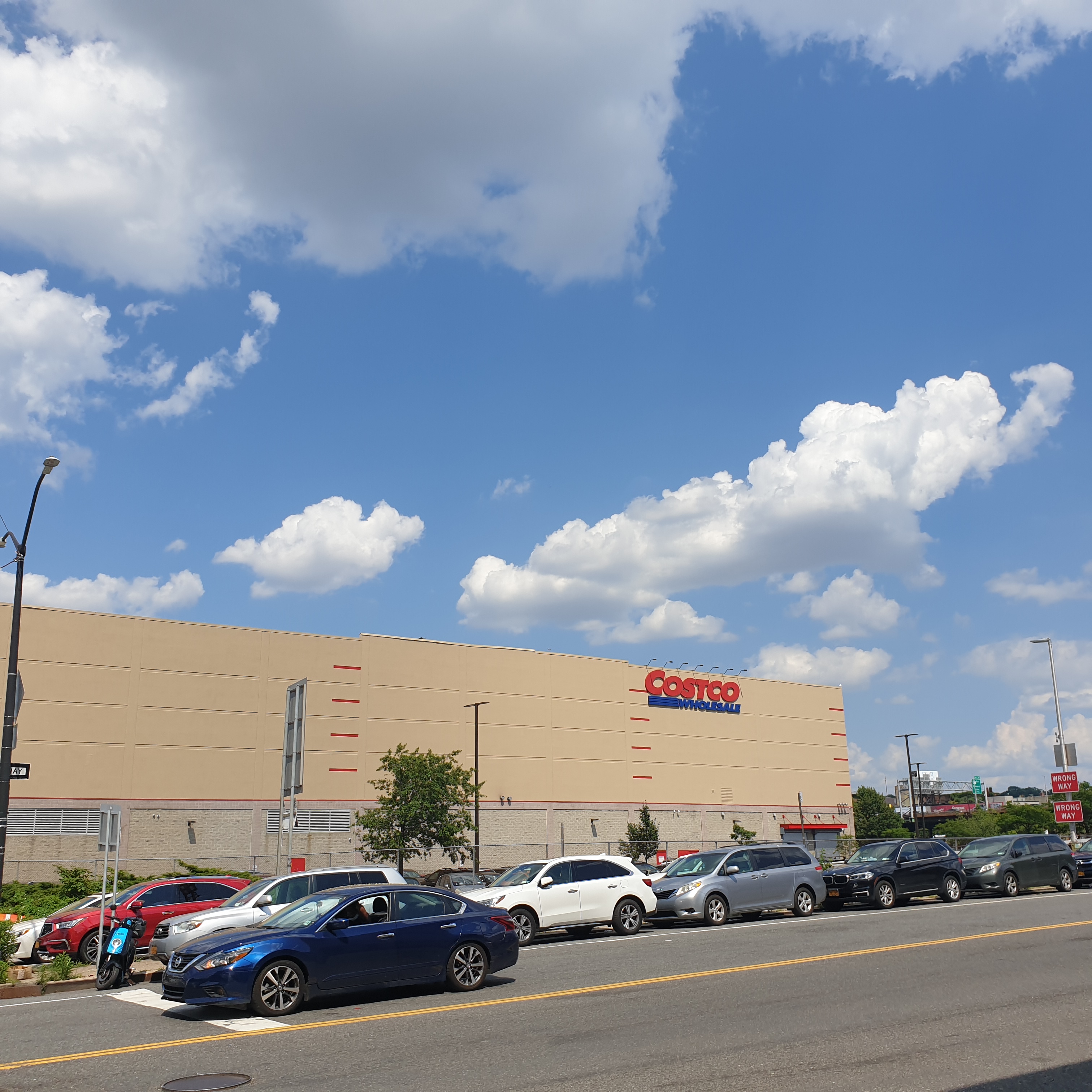 A street with one blue car driving on it and a number of cars parked on it, in front of a large Costco store with a long beige wall. There is a blue sky with fluffy white clouds.