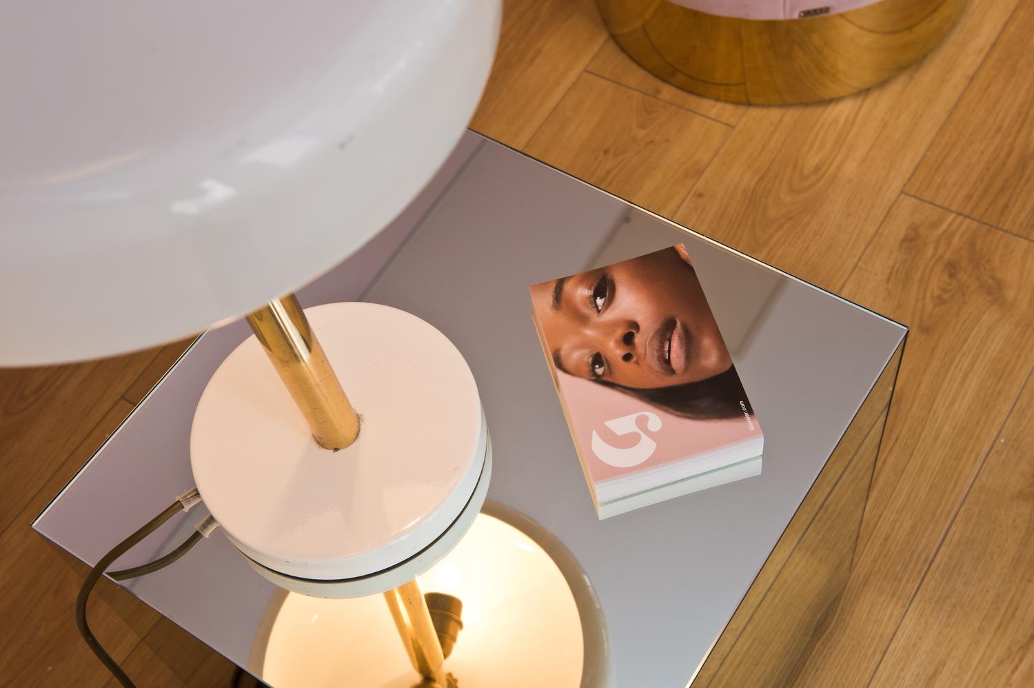 Glossier brand booklet with model’s face placed on mirrored side table next to a lamp.