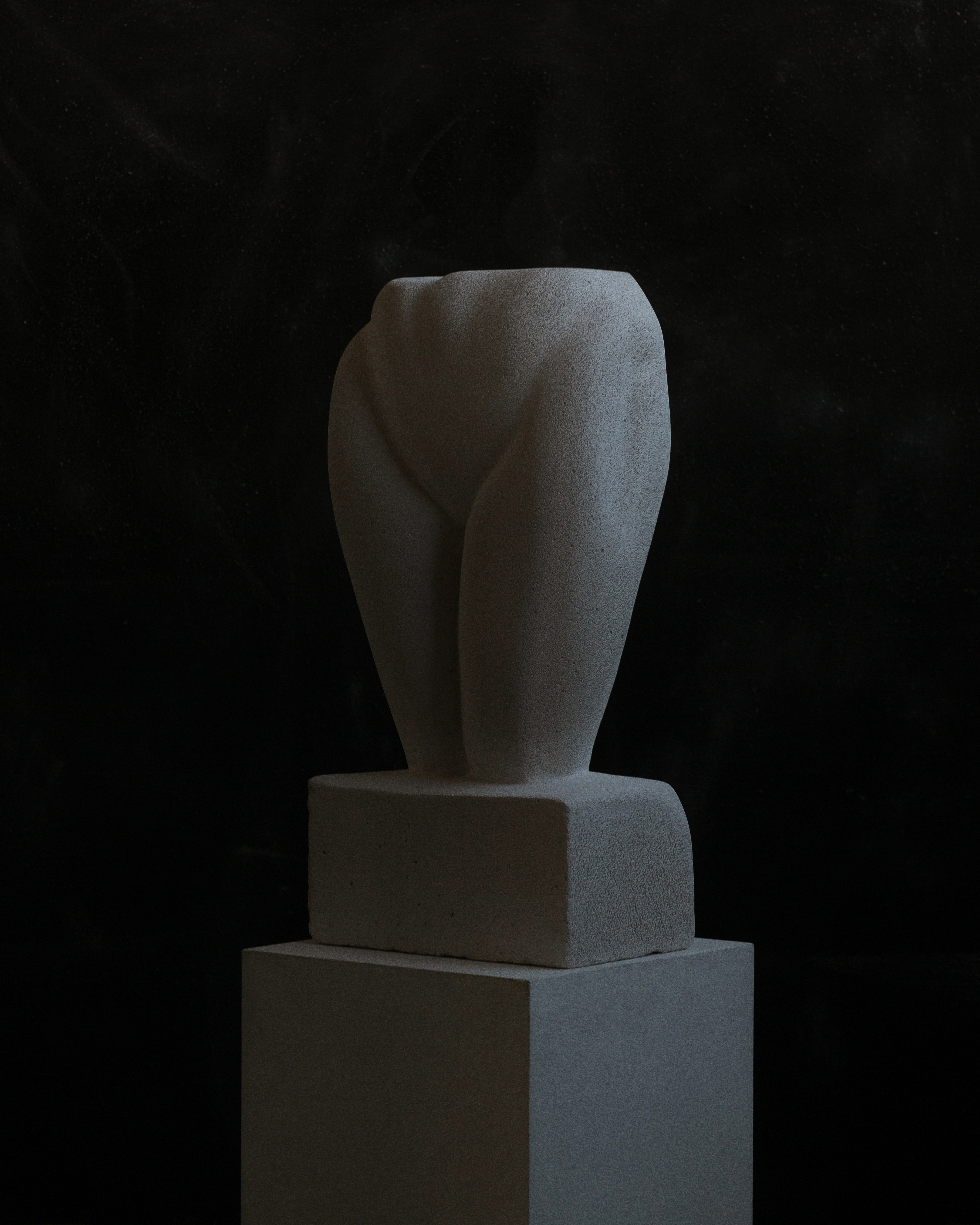 sculpture of a woman's hips and thighs at an angle carved out of concrete and stone sitting on top of a white wooden plinth against a black textured background