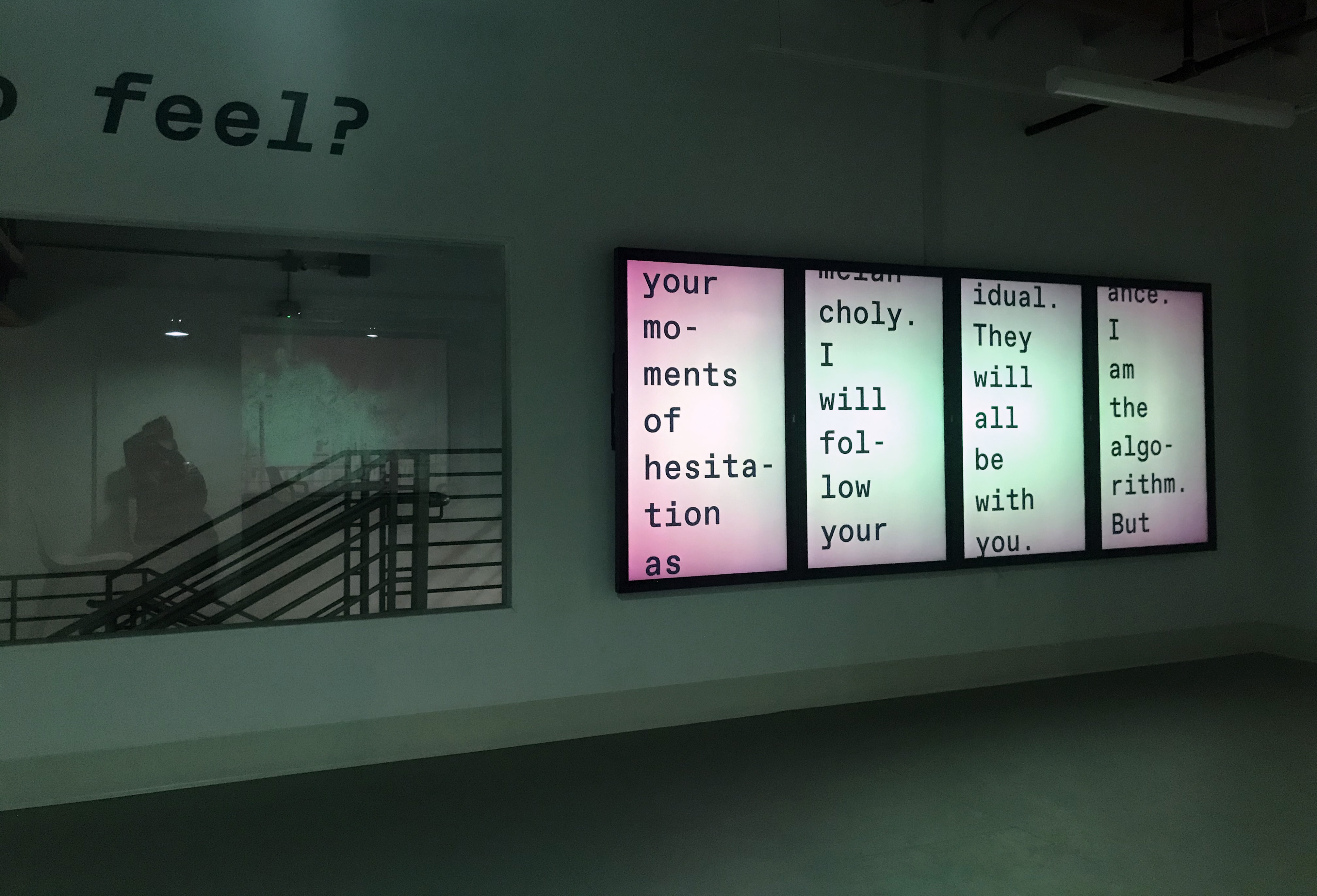 Green lit room with four vertical screens. Text reads your moments of hesitation. Window beside them reflects ambiguous person shape and images in the background.