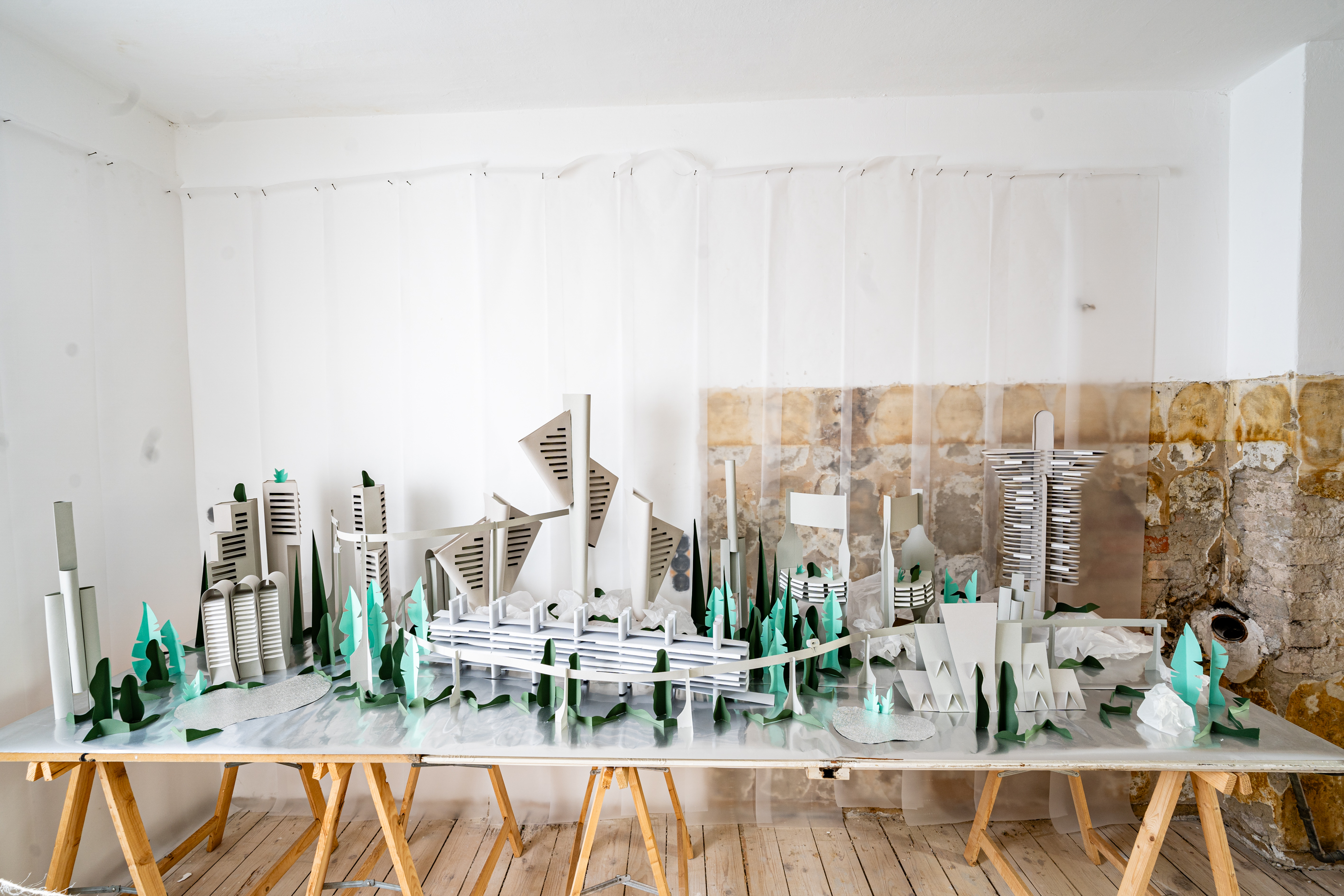 Bild 1 zu Neometropolis: Leichte Aufsicht auf das Papier Modell, welches auf einer 2 Meter langen spiegelnden Tischplatte platziert ist, die auf B&ouml;cken liegt. Das graue Papiermodell zeigt hohe, sowie niedrige Geb&auml;ude, die alle auf geometrischen Formen basieren und von langen schmalen Fensterpartien durchzogen sind. Zwischen den einzelnen Geb&auml;uden sind gr&uuml;ne Modellb&auml;ume platziert.