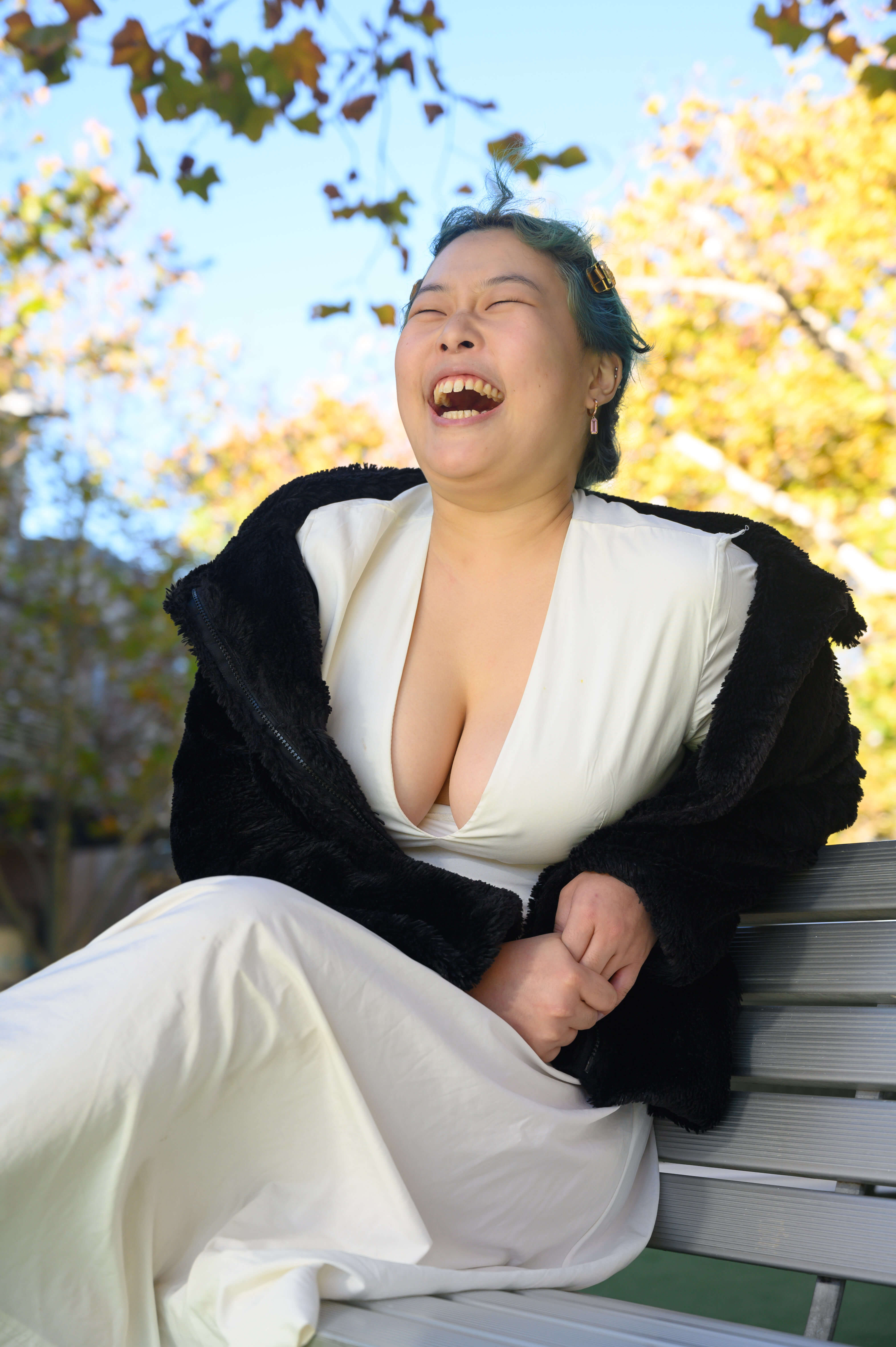 Lydia, a light skinned Korean woman with turquoise hair, is poised mid-laugh. She clasps her hands by her waist as she sits on a bench.