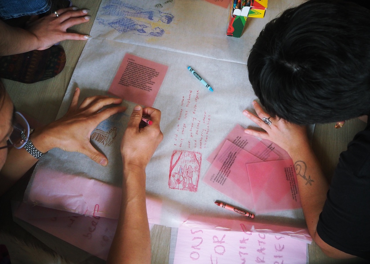 Several people’s hands travel to write and draw across quietly lit chart paper. A pile of transparent pink notes slip under one of the participant’s wrists while an opaque pink note grazes the tip of another participant's hand bracing as they mark the chart paper.
