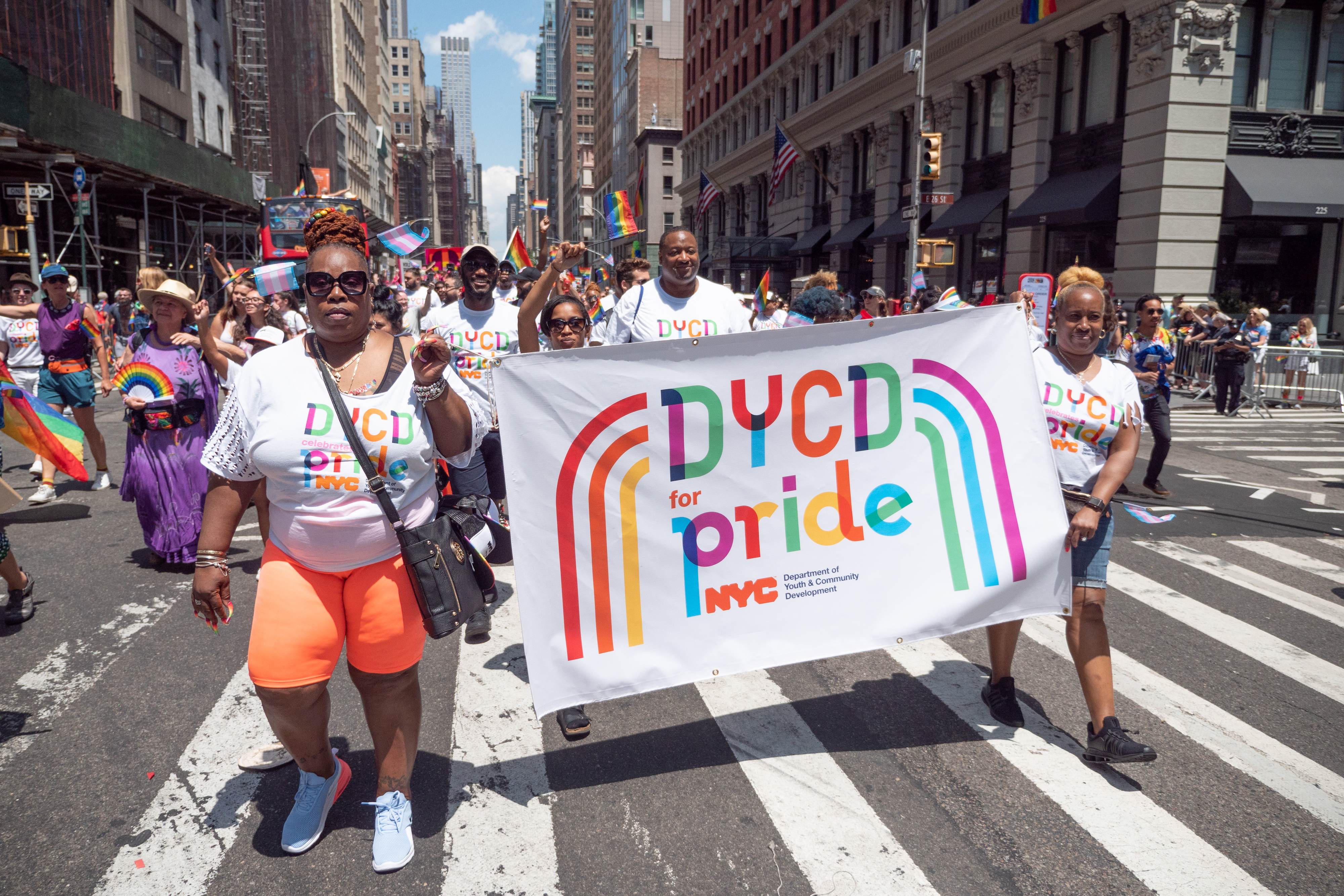 DYCD employees marching the 2019 NYC Pride parade wearing uniform branded white shirts with the DYCD celebrates Pride rainbow graphic. Two DYCD employees march holding a white banner that displays the DYCD for pride rainbow graphic.