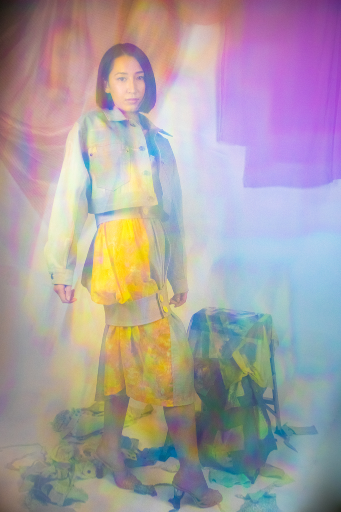 A woman in a yellow skirt and denim jacket stands in front of a colorful fashion collection, with a white background and a rainbow of light reflecting through the image. Ethereal and visually striking.