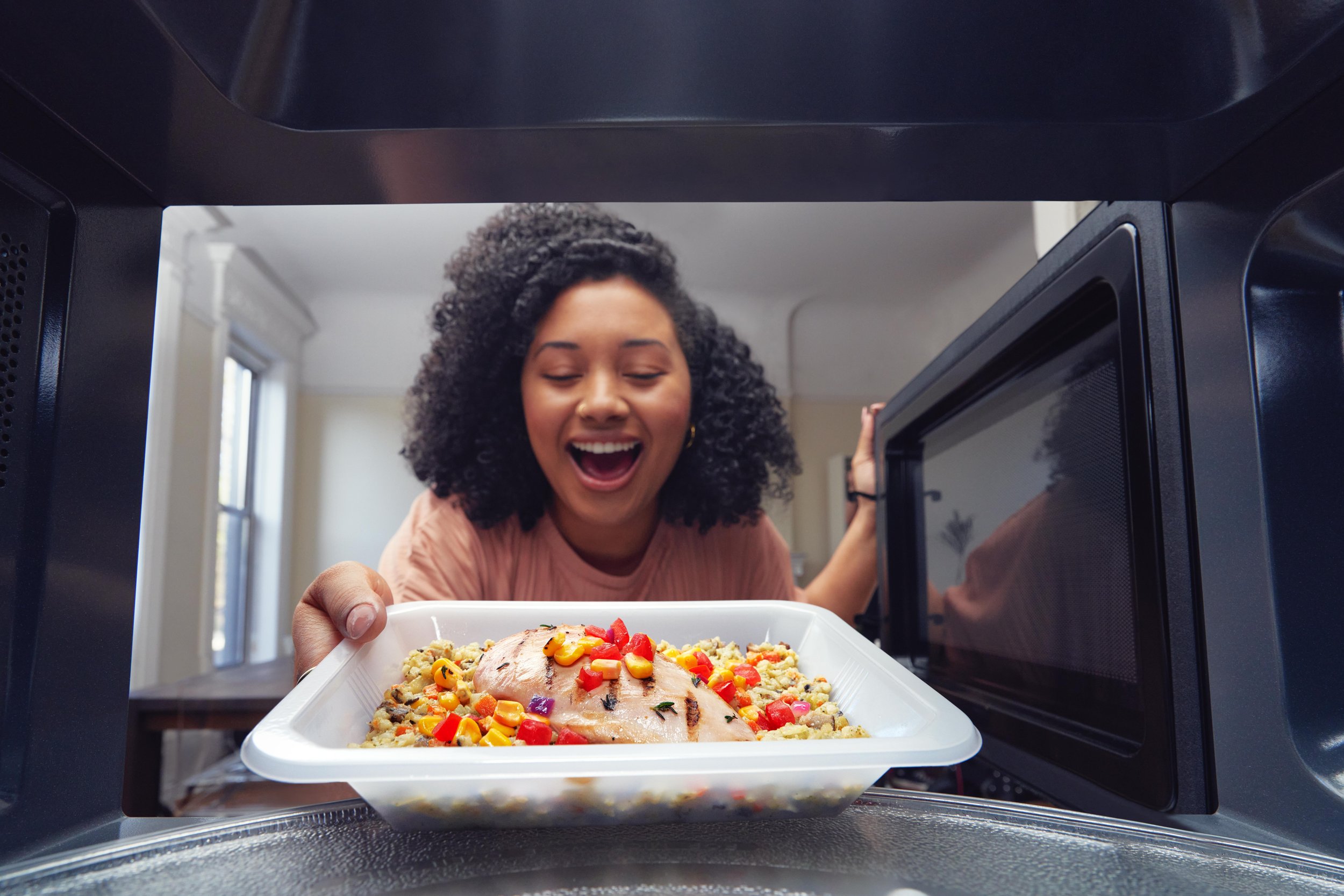 A woman pulls a freshly-cooked meal from a microwave smiling; shot from the perspective of inside the microwave looking out.