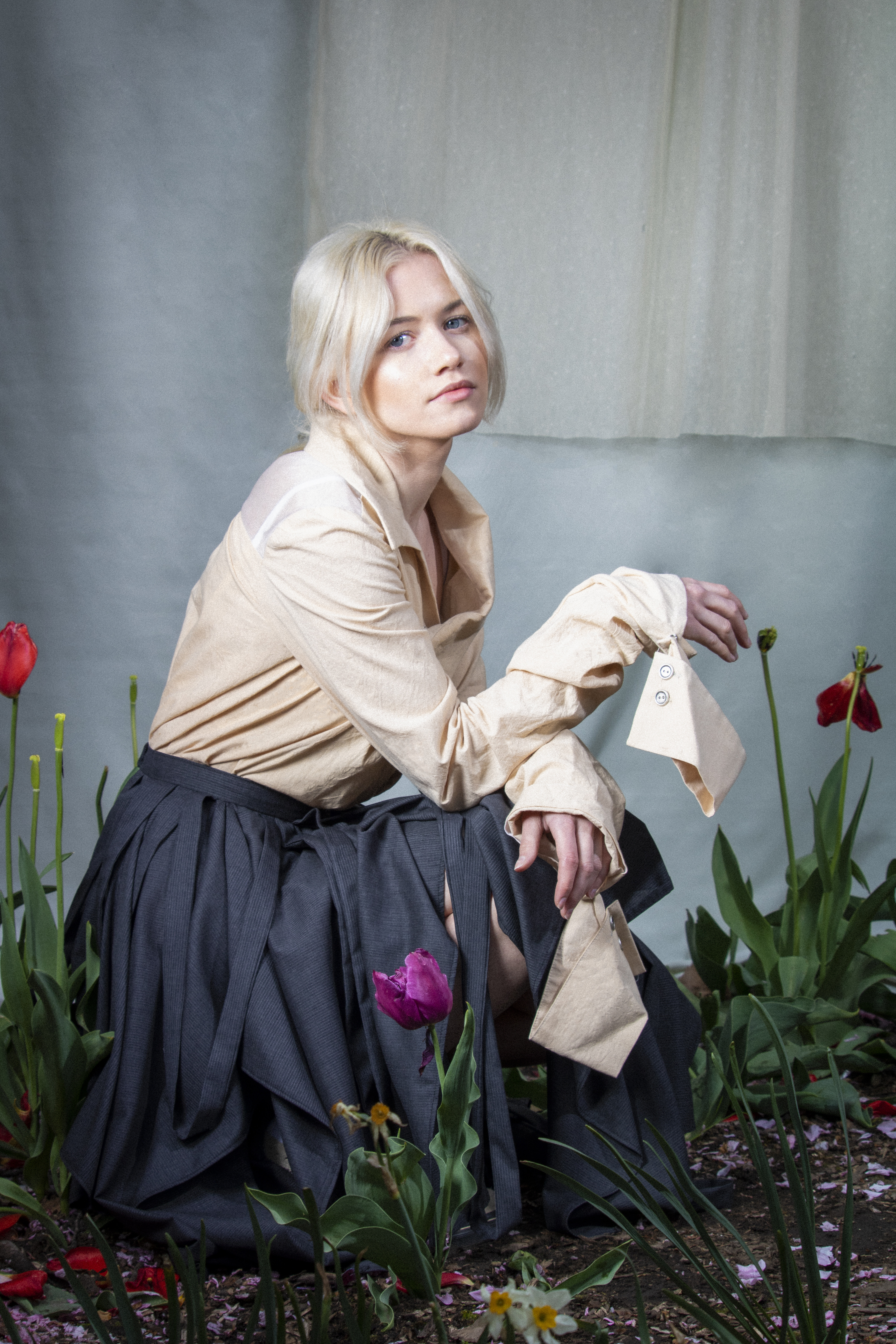 Amidst a vast sea of tulips, a blond woman, Emailia Kaplan, kneels gracefully, exuding an experimental fashion narrative. Her beige blouse and dark pleated skirt contrast with the vibrant floral backdrop, while her neutral expression adds an air of quiet introspection.