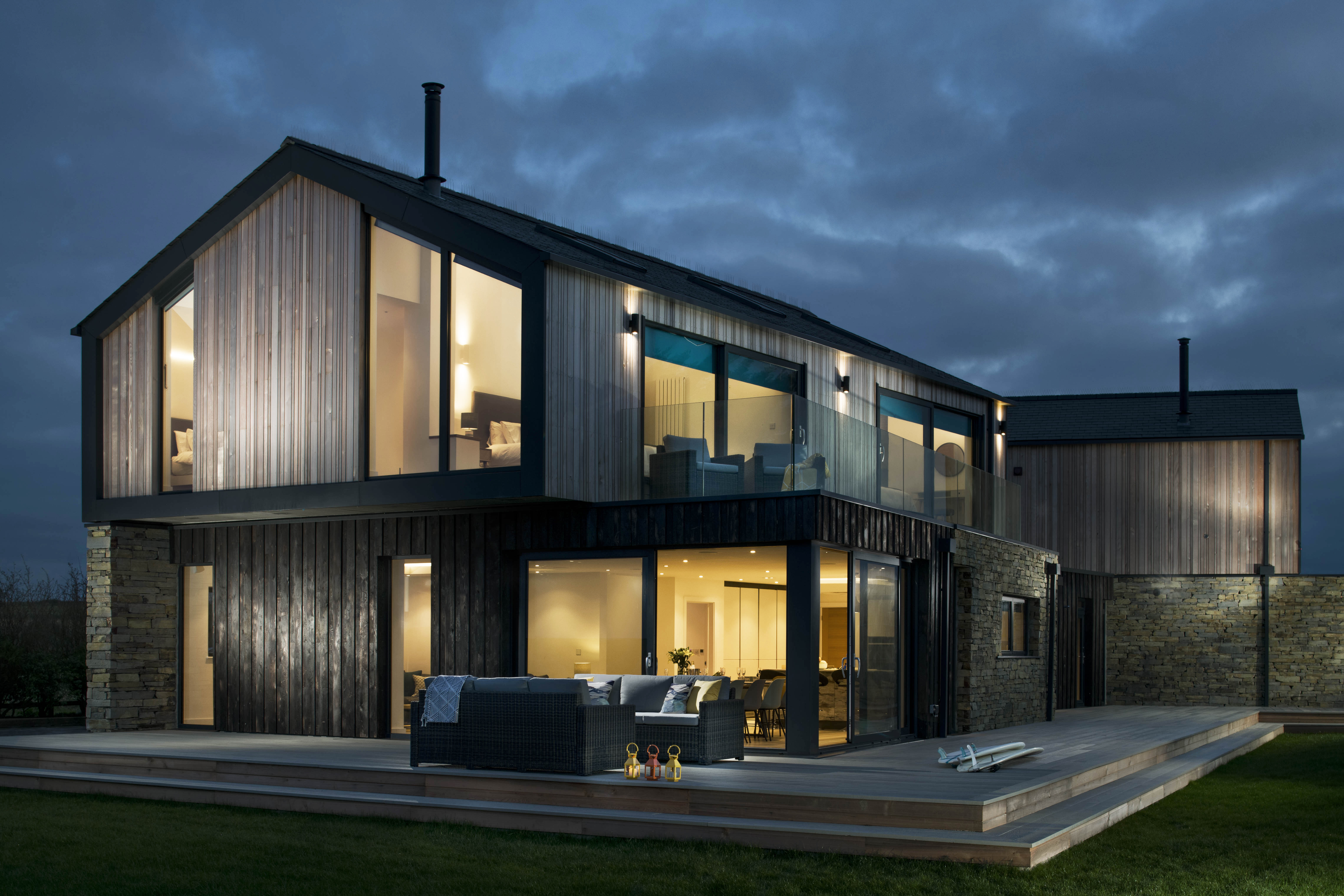 House exterior at dusk. Timber and stone material on facade. Rattan furniture on raised timber patio. Dimly lit interior. 