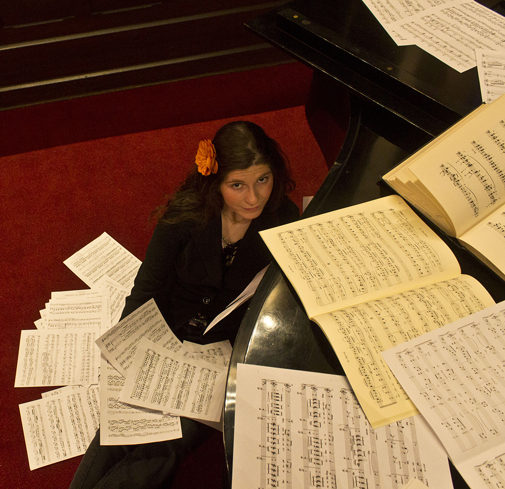 A person sits on a carpeted floor and leans against the piano. There are sheet music scattered all over the piano and around the person.