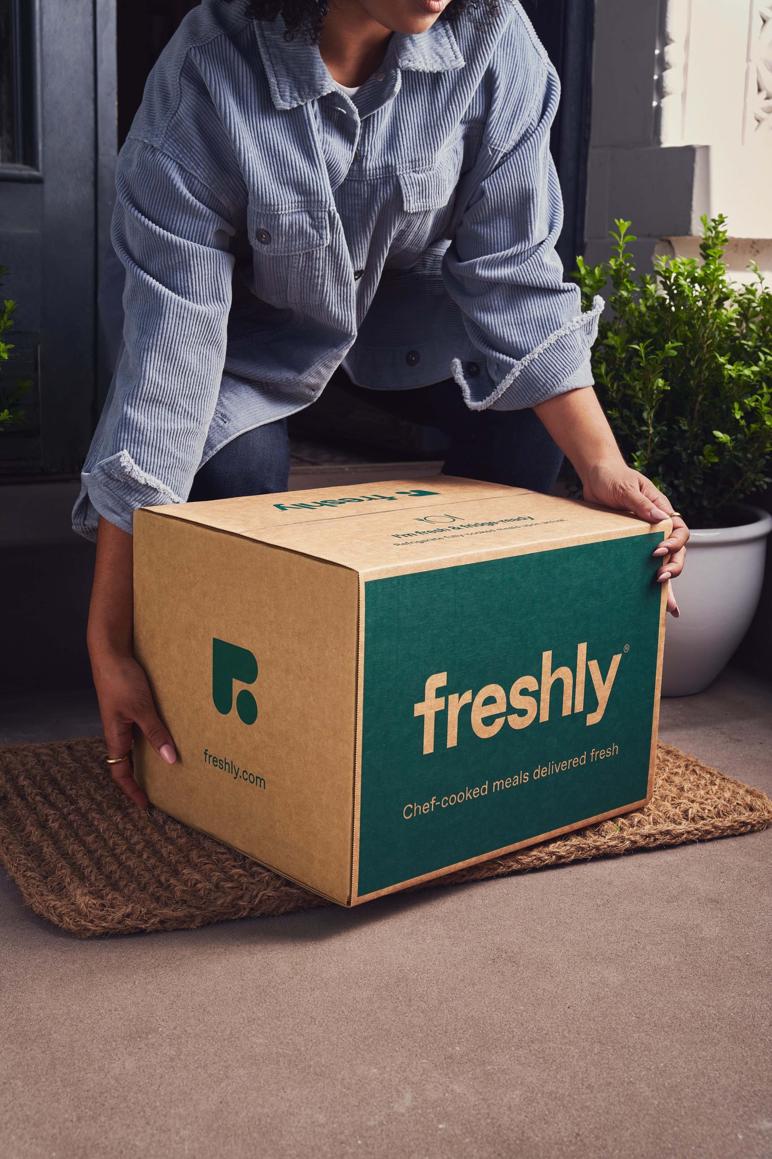 A woman in a blue jacket kneels down to pick up a just-delivered box of Freshly meals from her doorstep.