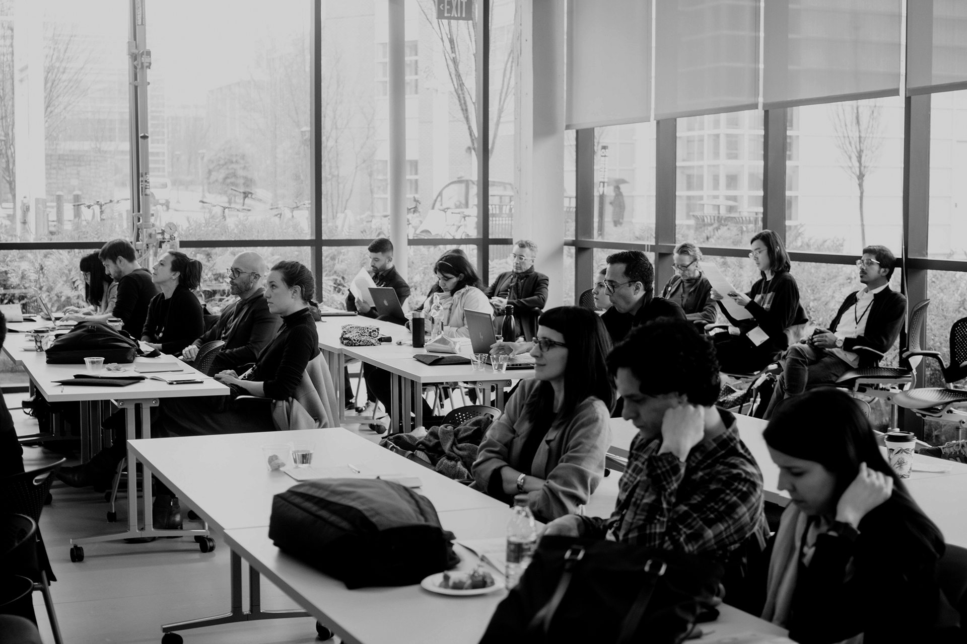 Seventeen people sitting in a group listening to a man give a lecture. Lecturer has a laptop on the table infront of him.
