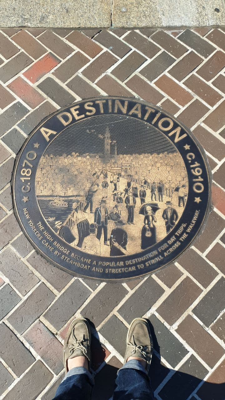 A pair of feet in front of a circular plaque on the ground. It reads "1870 A Destination c.1910" and "The High Bridge became a popular destination for day trips. New Yorkers came by steamboat and streetcar to stroll across the walkway."