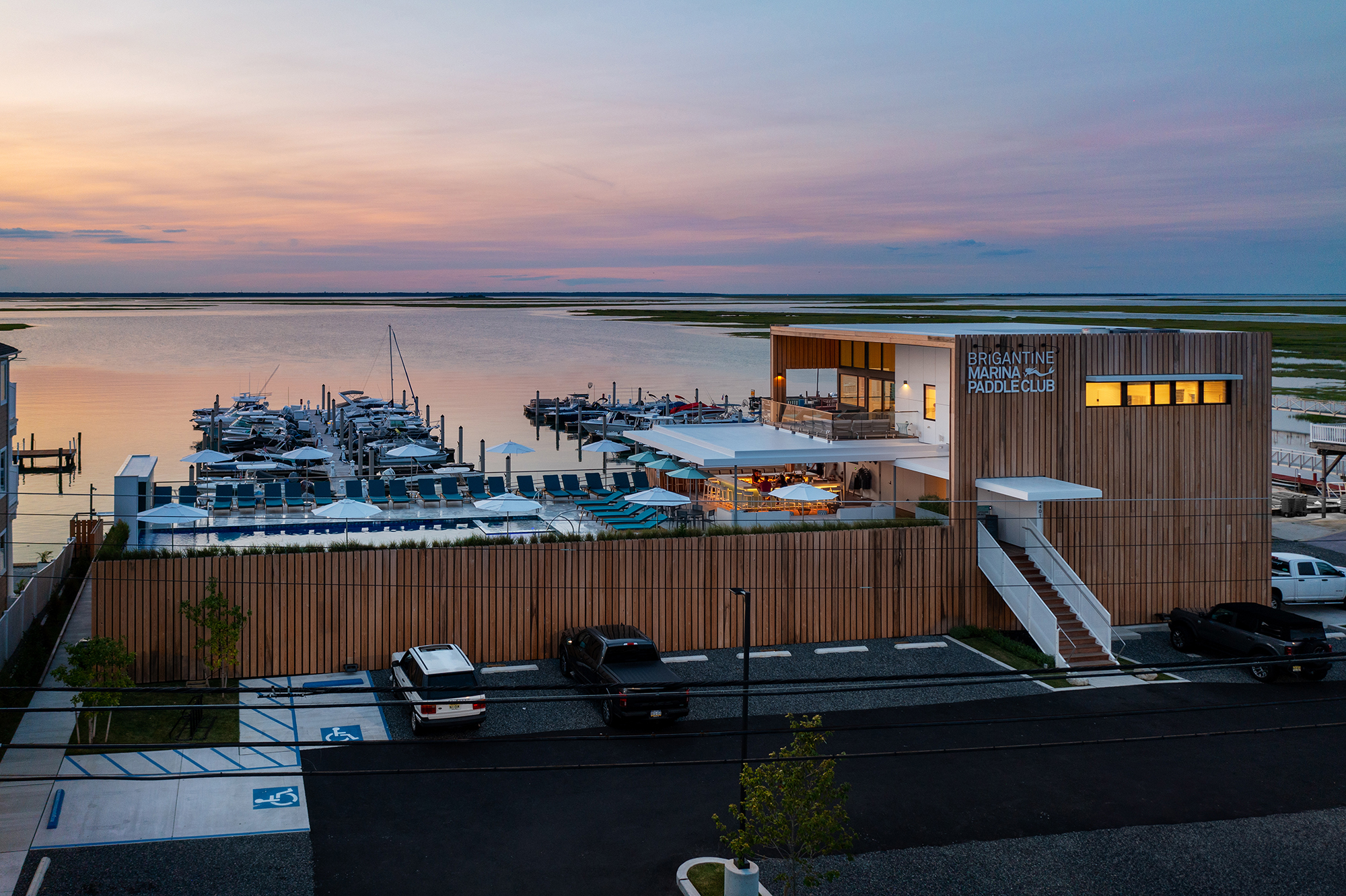 Exterior photograph of the Brigantine Marina and Paddle Club at sunset, showing the building with Cypress wood cladding, an elevated pool deck overlooking the marina, and docked boats on the bay waterfront.