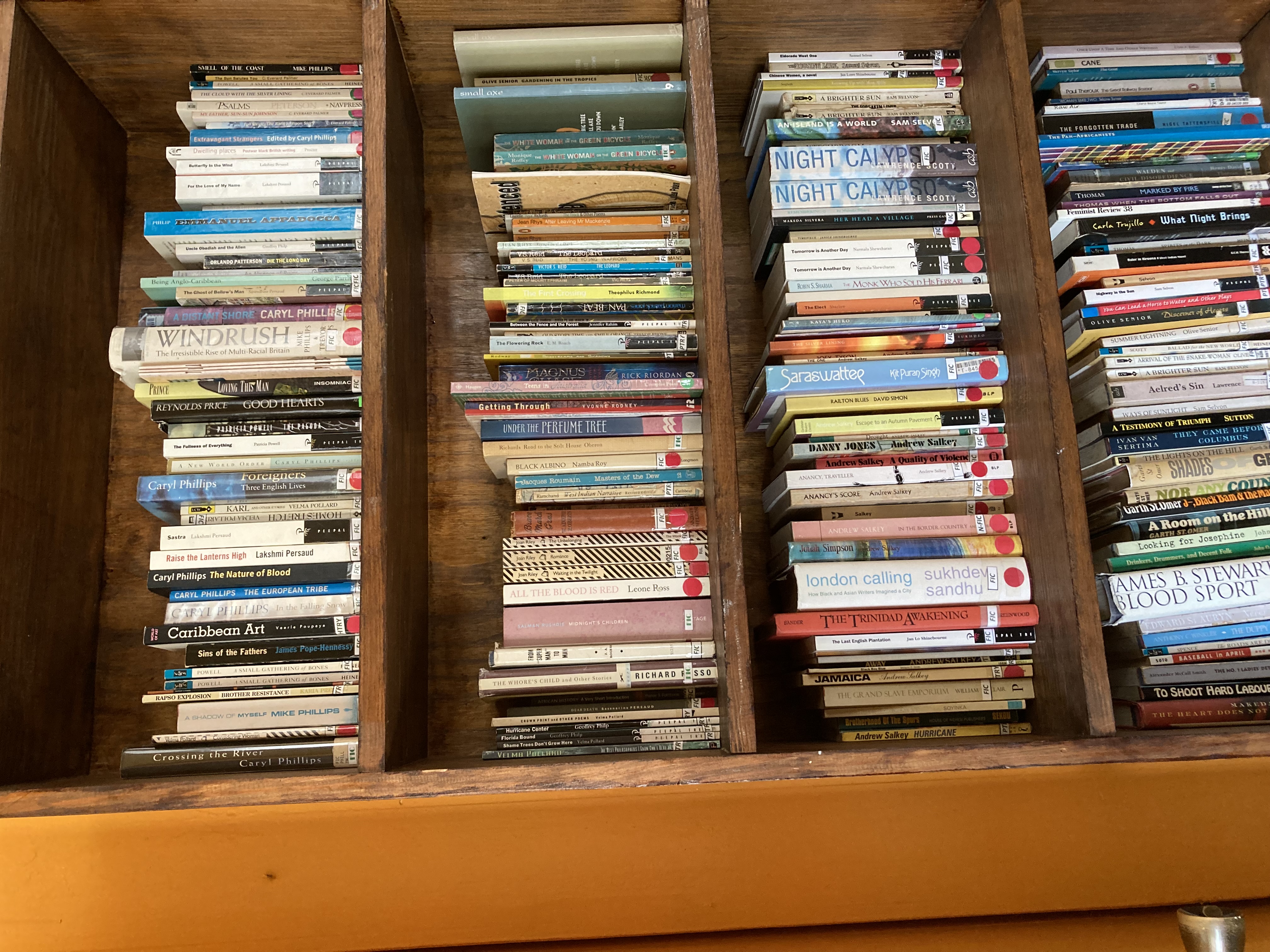 a wooden bookshelf with 4 shelves, most of the titles are too small to read but some of the larger text titles on the spines of the books say "windrush" and "caribbean art" and "night calypso" and "the trinidad awakening"