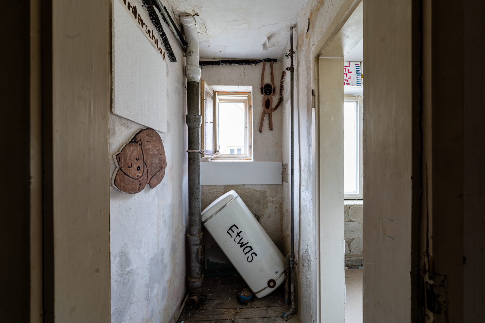 Bild 1: Zwischen schwarz und wei&szlig;: Das Bild zeigt einen schmalen Raum, ein ehemaliges Klo mit einem kleinen ge&ouml;ffneten Fenster auf der gegen&uuml;berliegenden Wand. Das Klo wurde entfernt, daf&uuml;r liegt schr&auml;g an der gegen&uuml;berliegenden Wand ein Boiler unterhalb des kleinen Fensters auf dem das Wort "Etwas" geschrieben steht. Links an der Wand ist ein schlafender brauner B&auml;r gemalt, neben dem Fenster h&auml;ngt ein gemaltes &Auml;ffchen. Rechts geht es weiter in den n&auml;chsten Raum. 