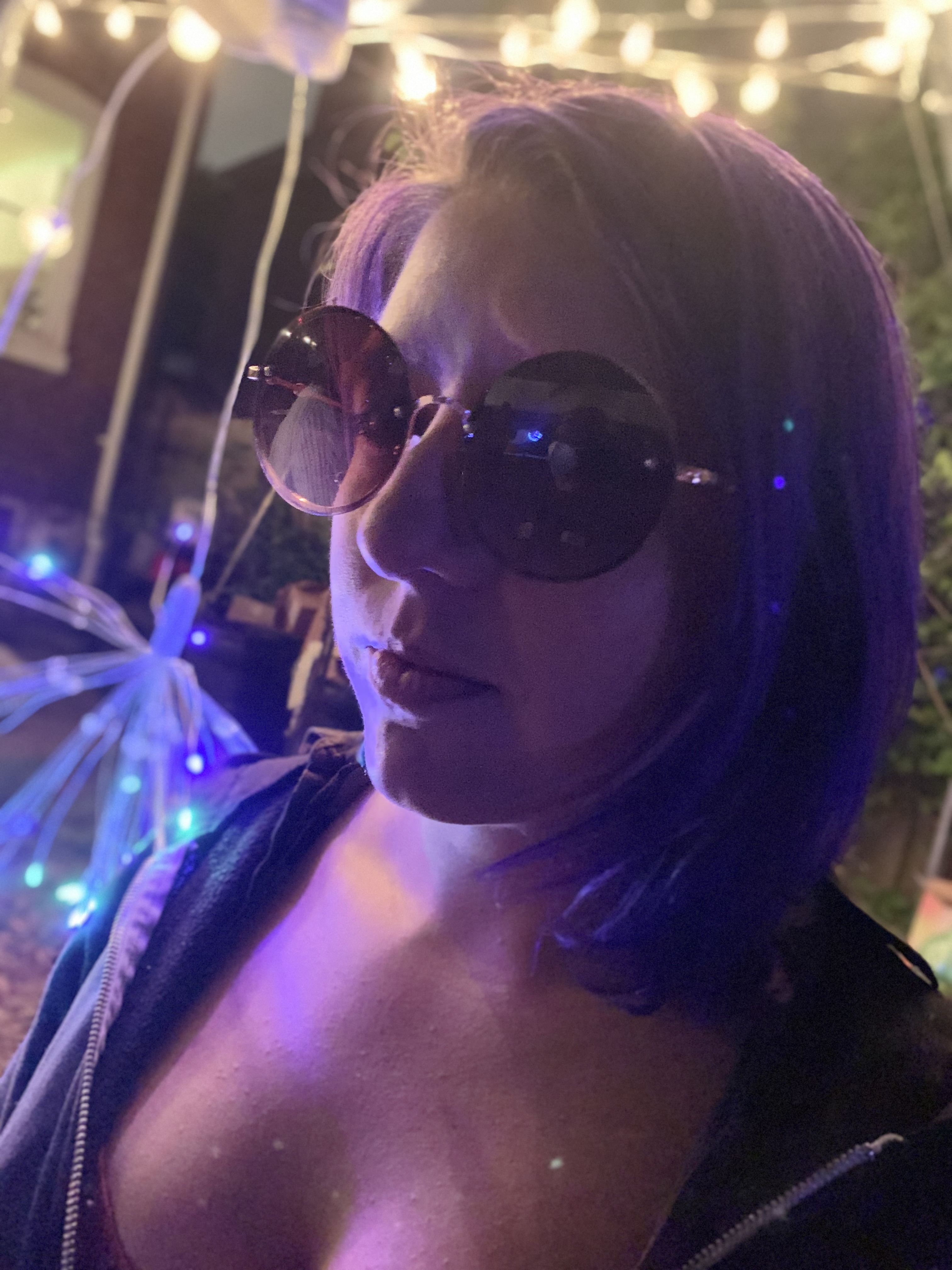 Portrait headshot of Tobin Ibershoff, who is looking slightly to the left and wearing large circular sunglasses and a zip up hoodie in a nighttime scene with lights, possibly a party.