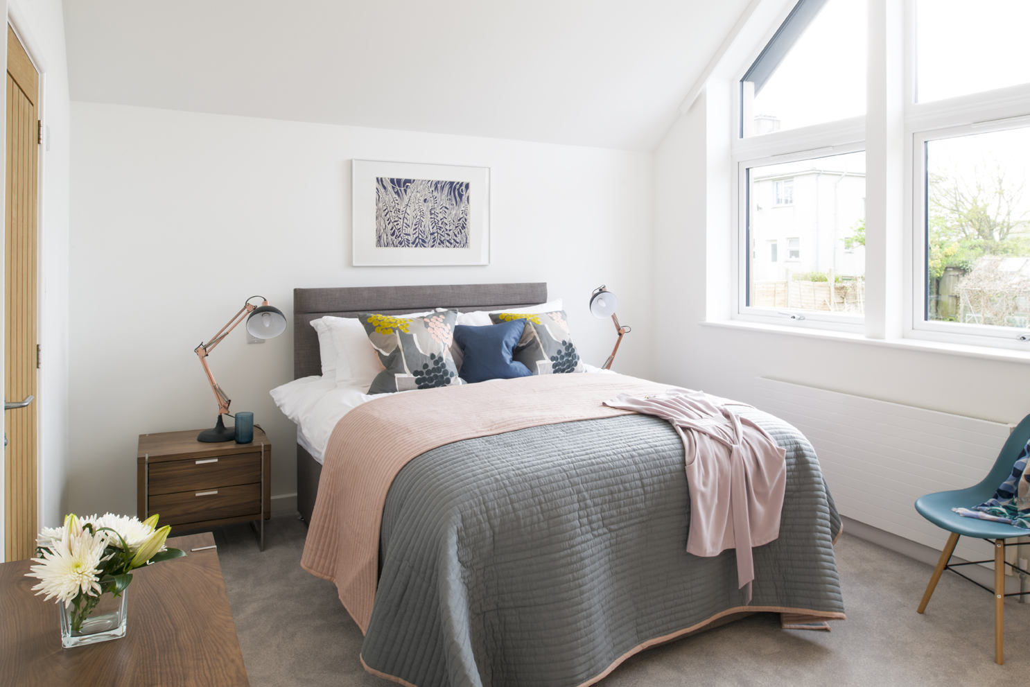 Modern bedroom with double bed with pink and grey bedspread. Teal chair in corner and wooden side tables with lamps.