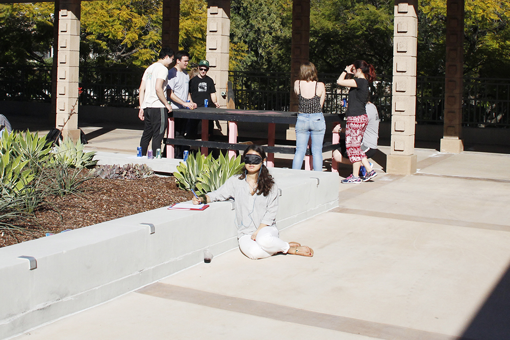 A blindfolded performer sits on the ground and, on her right side, leans against the cemented edge of a raised garden bed. The performer rests her right arm on the same edge and writes on a writing pad. A group of six people stand around a table in the background.