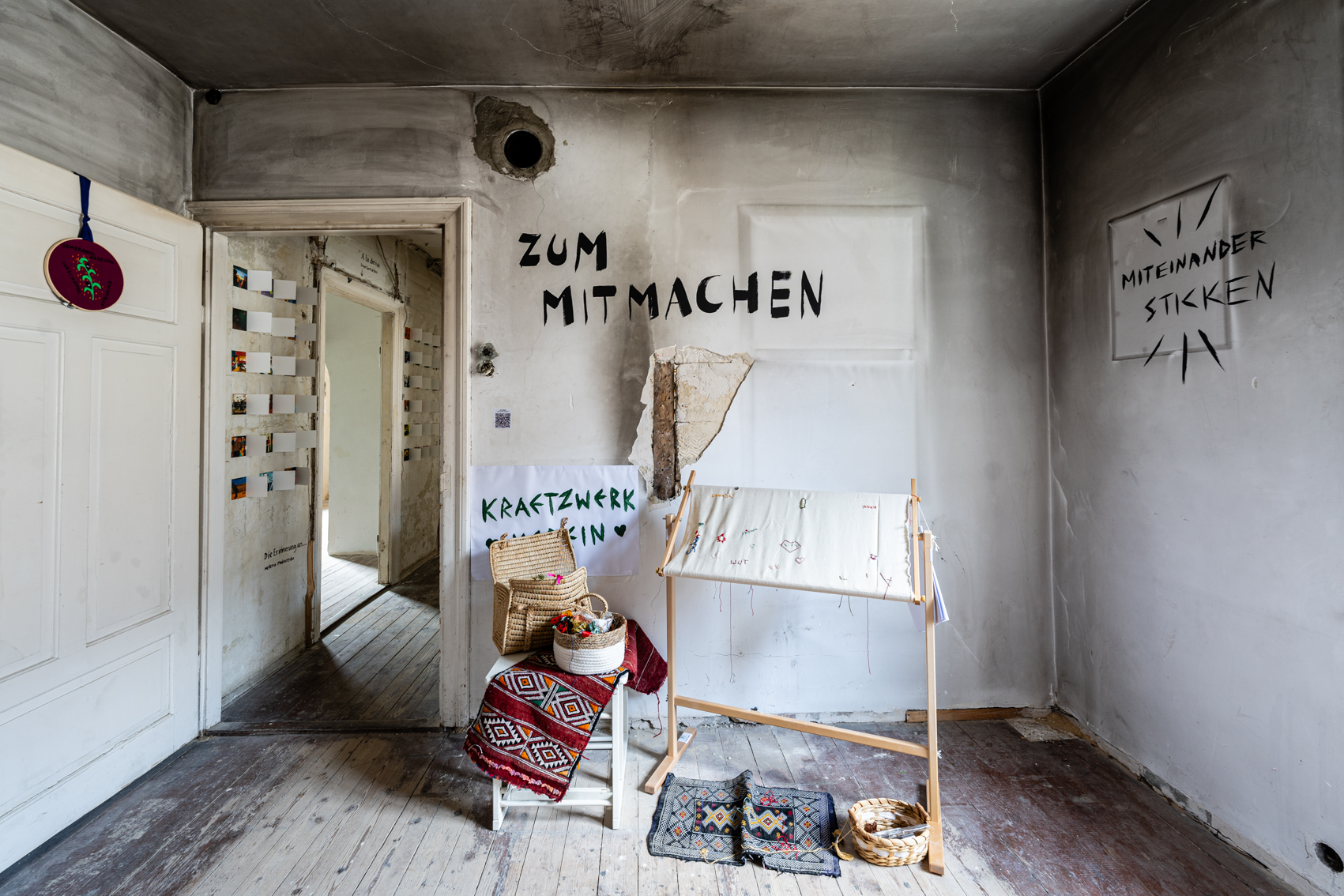 Bild 2: Wand rechts der Eingangst&uuml;r. Ein Stickrahmen steht vor der Wand. Am Boden liegt ein kleiner Teppich. Links des Rahmens ist ein Korb mit Garn zu sehen. An der Wand steht in schwarzer Handschrift : "Zum Mitmachen".