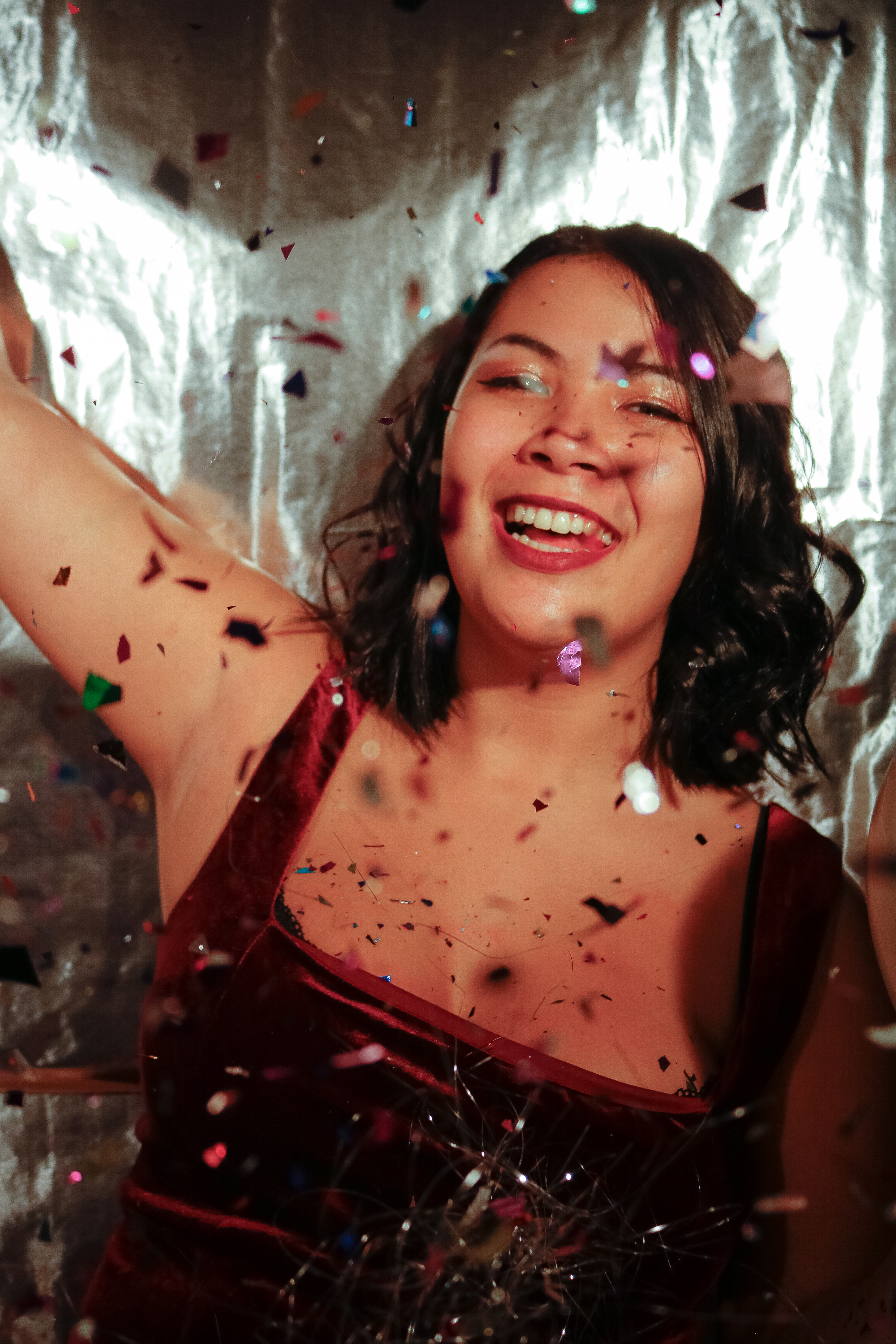 New Year's Eve-inspired image of an Asian woman in a red velvet dress, joyfully throwing glitter against a silver foil backdrop. Festive, dynamic, and celebratory.