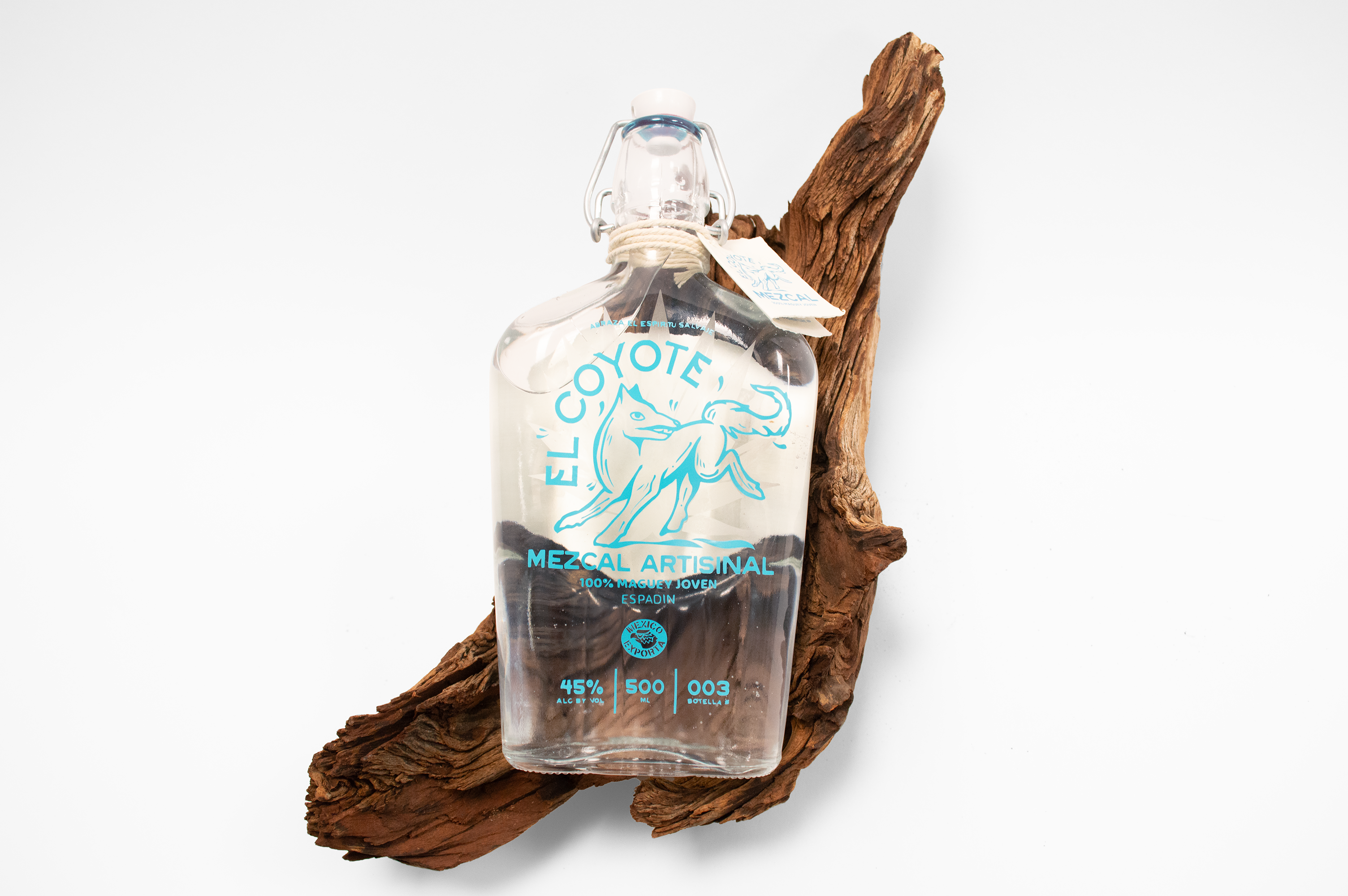 El Coyote Mezcal bottle laying on top of a rugged piece of drift wood on a white studio background.