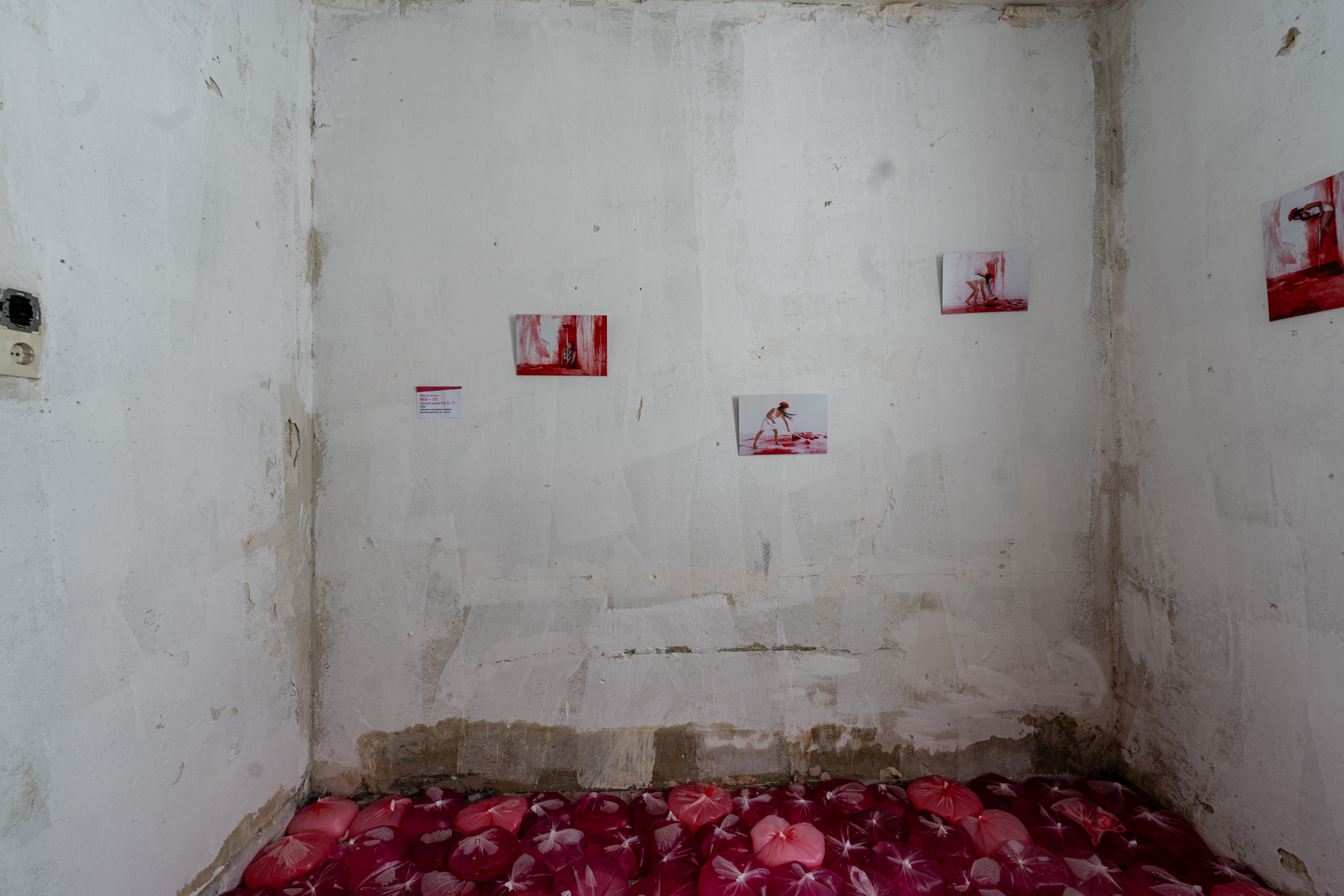 Bild 2 zu RED: Ansicht der anderen Seite des quadratischen wei&szlig;en Raumes. Die Wand zeigt ebenso wie Bild 1 Fotografien, sowie Farbbeutel am Boden.