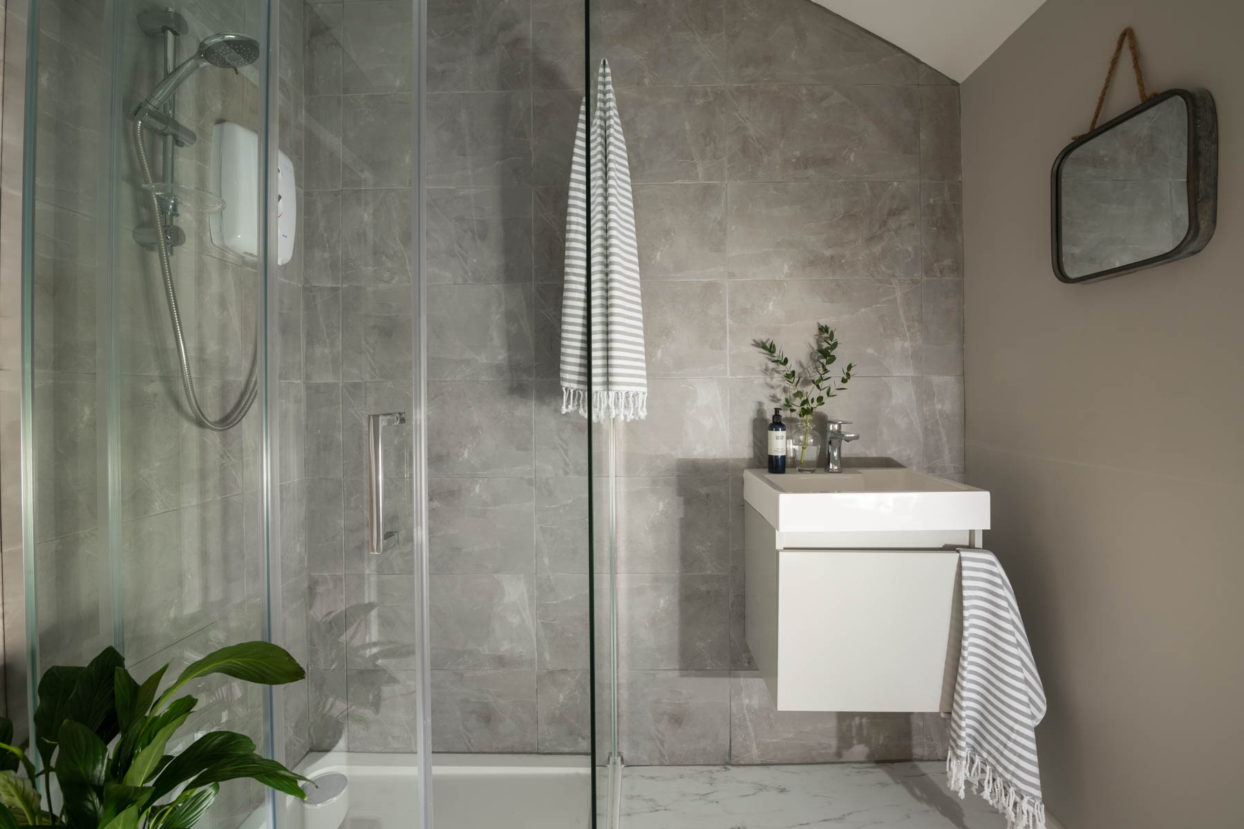 Grey tiled and painted modern bathroom, with white basin, mirror and shower with glass screen. House plant on marbled floor.