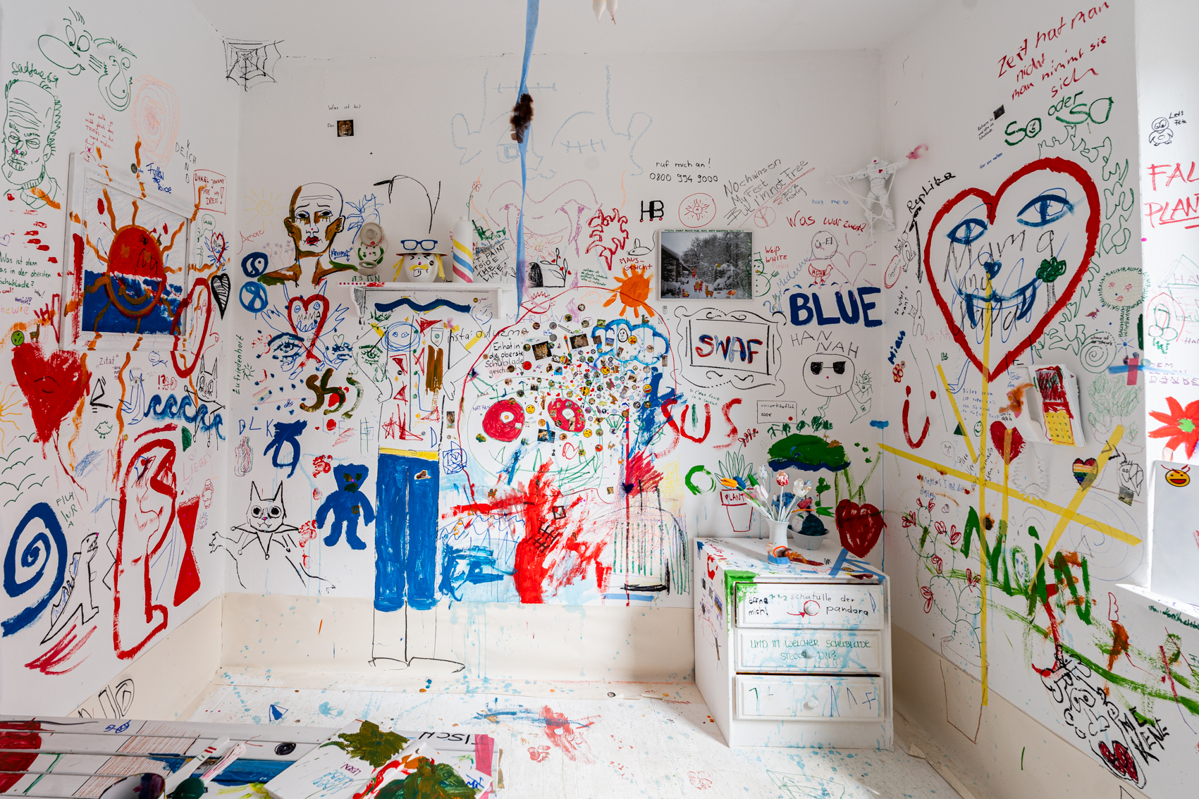 Bild 3 zu White Room: Im Raum stehend, rechts die Seite mit dem Fenster. Blick in den bunten Raum. An der Wand ist ein Regal befestigt mit wei&szlig;en Gegenst&auml;nden sowie rechts im Eck eine niedrige, bunt bemalte Kommode mit drei Schubladen.