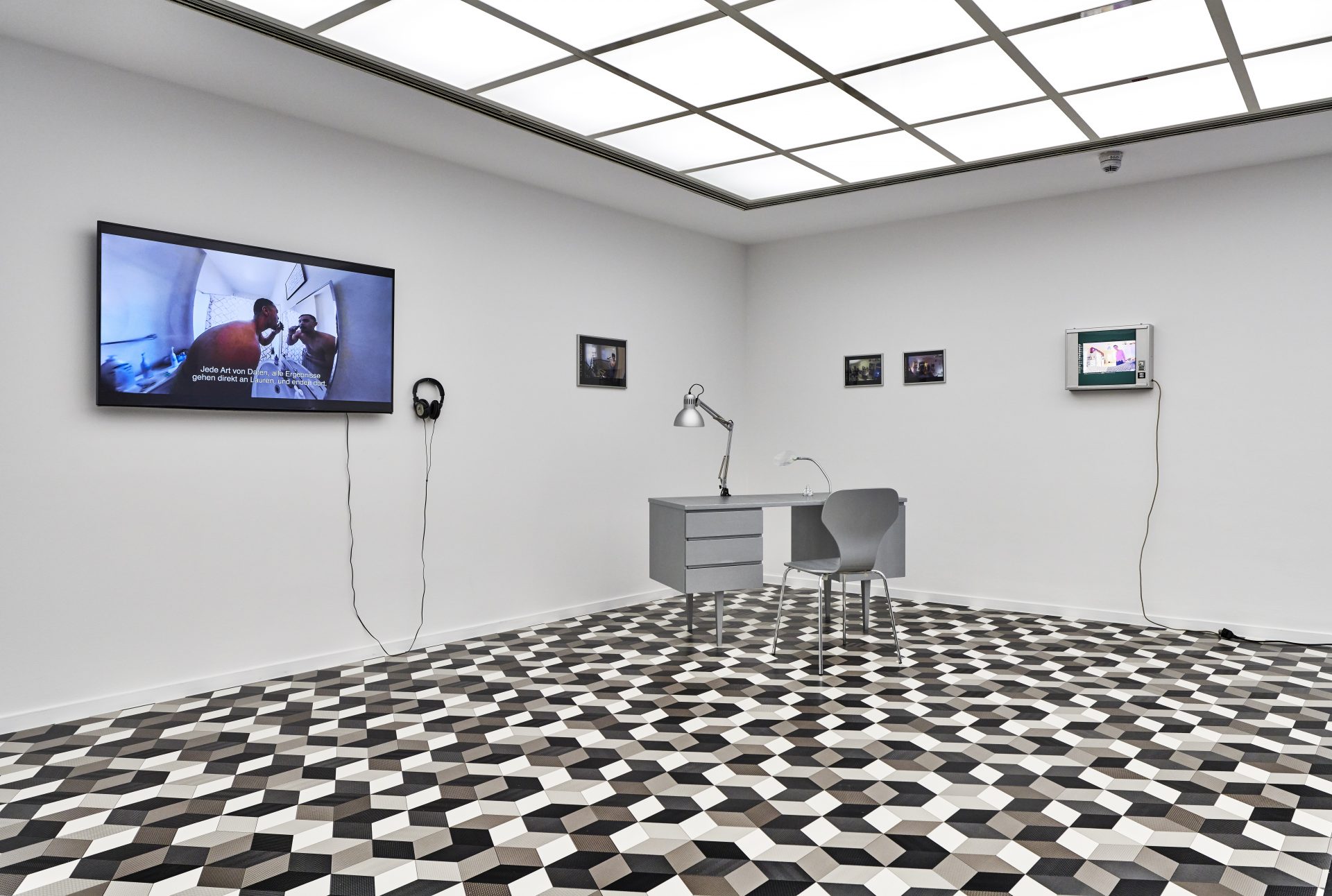 exhibition space with five screens on the wall, a video playing on each one and a gray desk with a chair and lap in the corner.