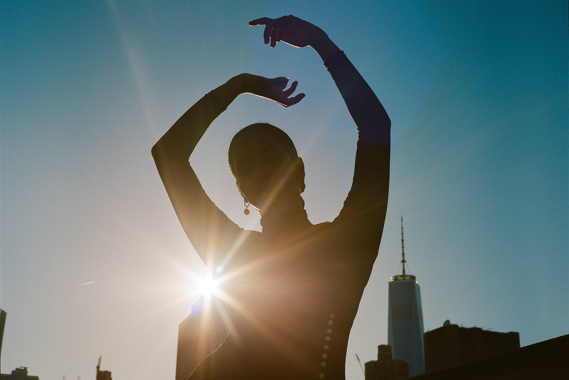A backlit model raises their hands above their head wearing Leigh NY, the background of the photo is New York City