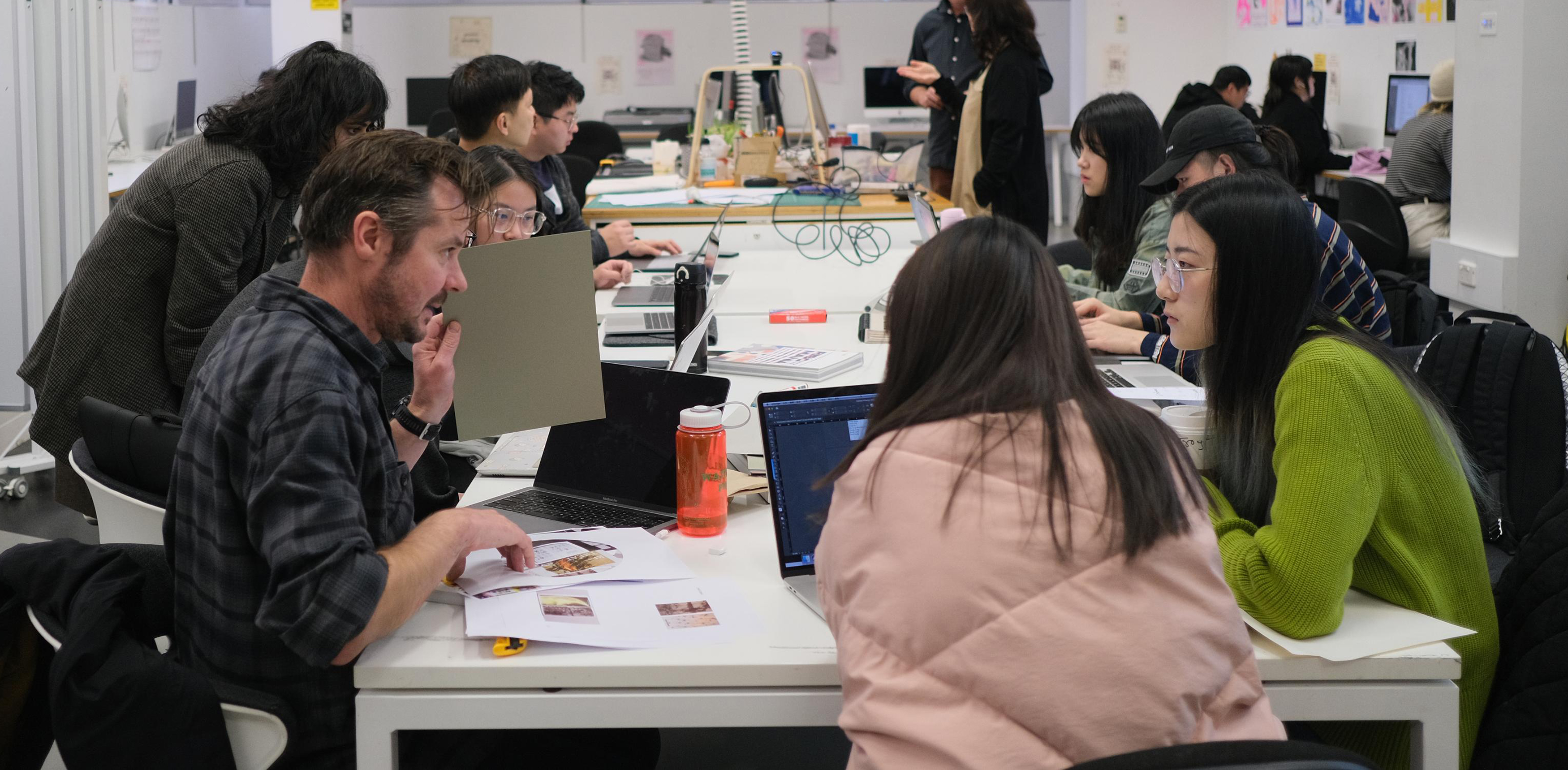 Lot of students are gathered in tables of studio and talking to each other.=