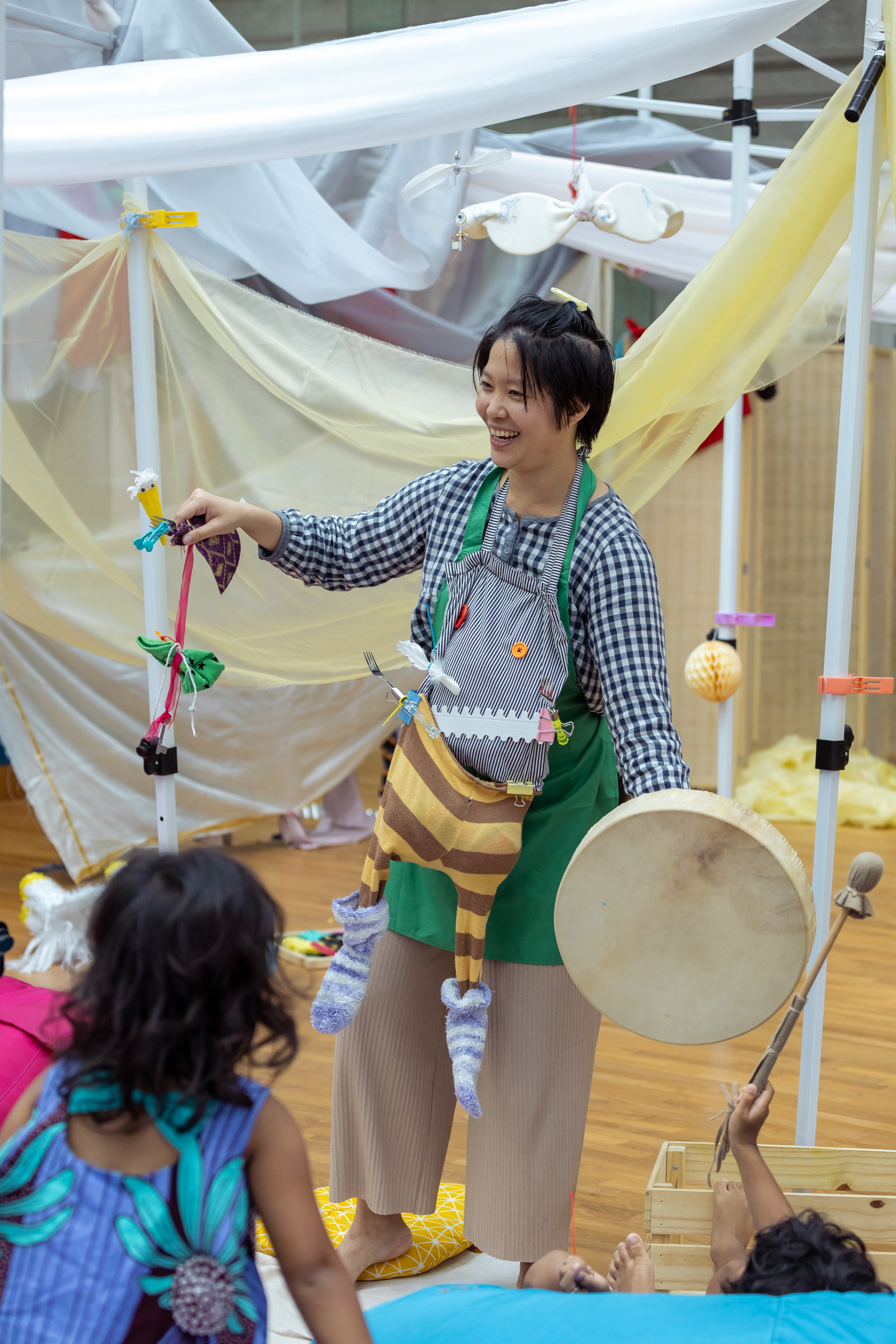 IMAGE DESCRIPTION: A performer in a quirky getup smiles at people outside of the frame as children watch on. Her hair clipped up by a clothes peg, she holds a hand drum in one hand, and a fork with googly eyes and dangling fabric in the other. Around her neck is a puppet made of a tote bag, buttons, office clips, and socks, and she has a green apron that contrasts with her blue-and-white gingham top.