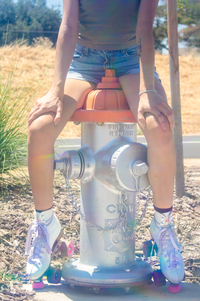 Bright, dreamy close-up of a girl sitting on a silver fire hydrant, wearing white and pink roller skates, Daisy Duke denim shorts, and a gray tank top. Only her lower half is visible, evoking a playful and nostalgic aesthetic.