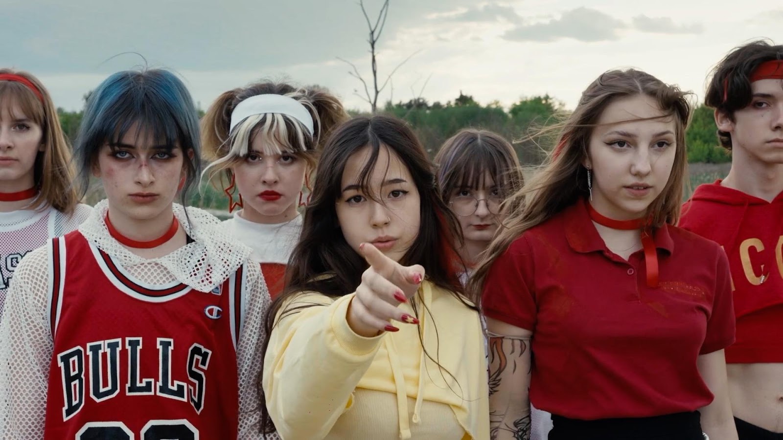 Video screenshot of a group of young people, looking sternly toward the viewer on an overcast day outside. They are mostly wearing reds and whites, except the centermost figure, who is wearing yellow, and has an outstretched hand, pointing at the viewer.