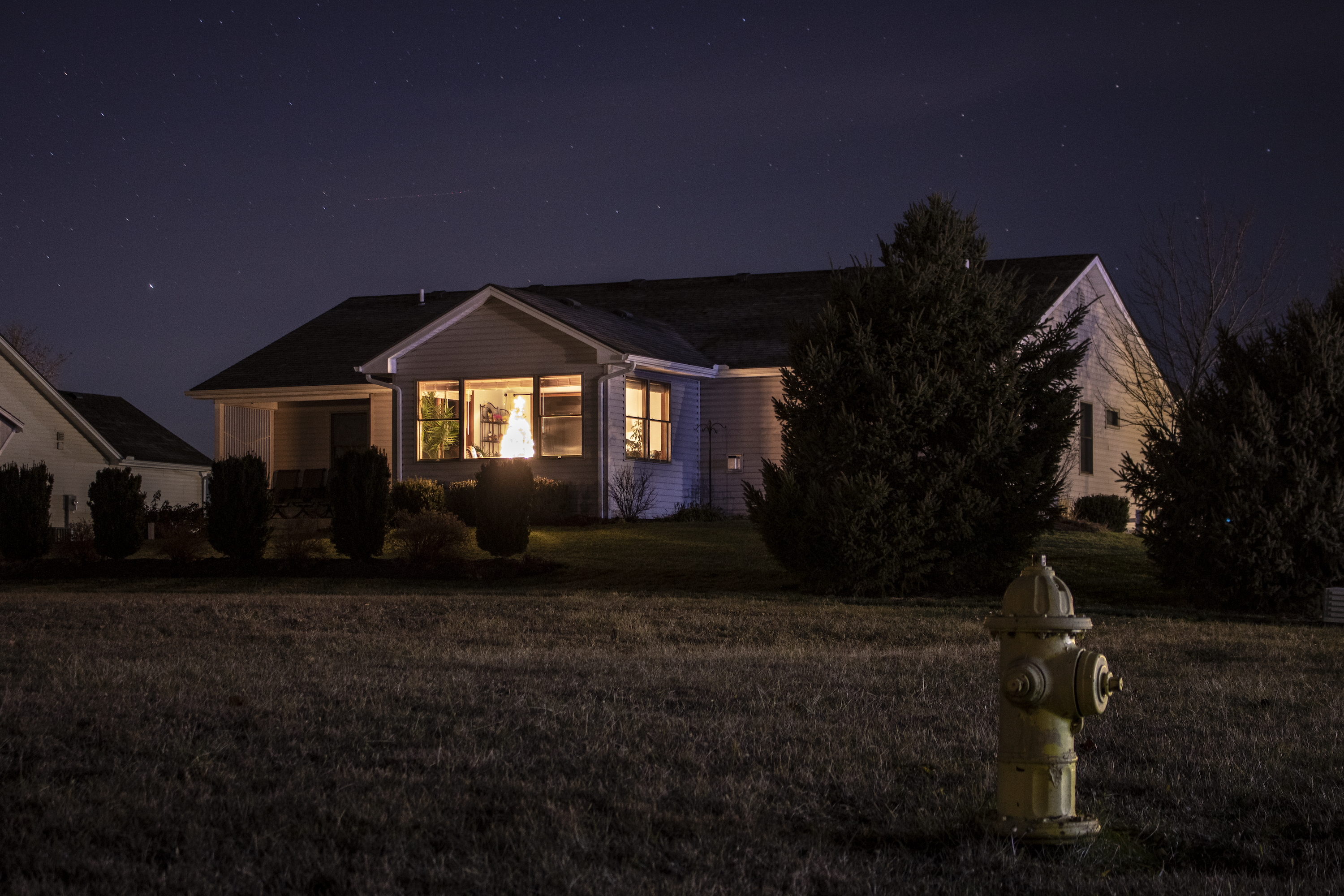 Nighttime scene of a house with a glowing Christmas tree inside, in Xenia, OH. Stars twinkle above, and a yellow fire hydrant stands in the foreground on the lawn.