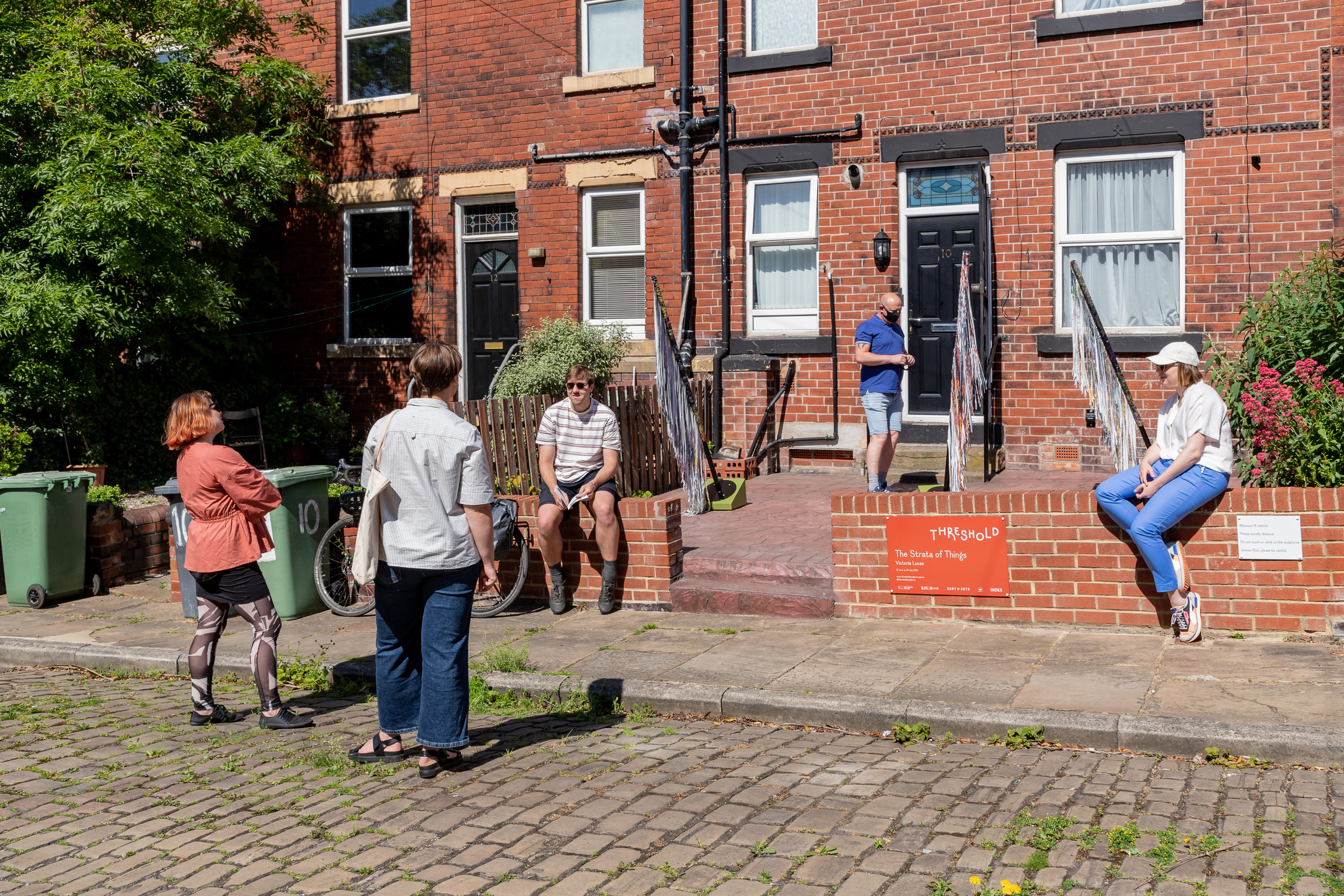 Visitors to the opening event sitting on the front wall, looking at the sculptures and chatting in the street.