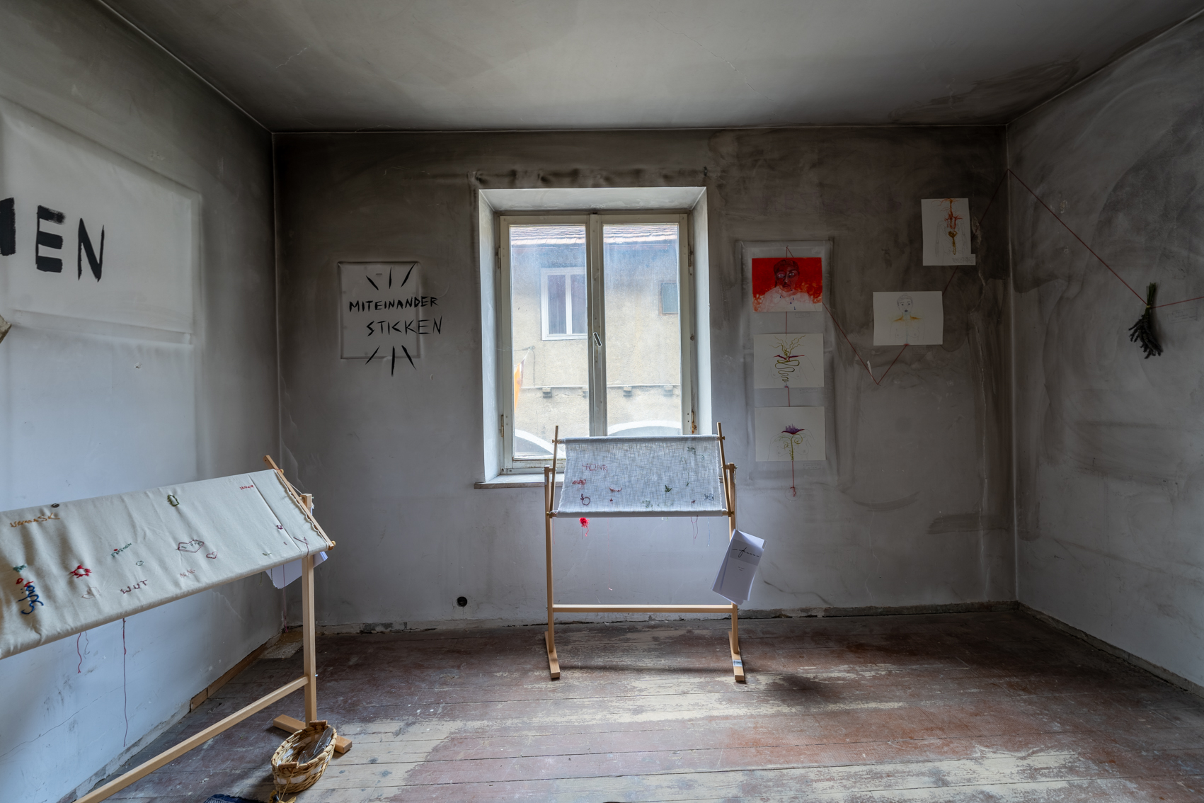 Bild 3 zu ImmerWiederNeuBeginnerin: Die Wand links der Eingangstür. Mittig ein Fenster, darunter der Stickrahmen von miteinander verwoben. Rechts des Fensters befinden sich noch fünf weitere Bilder. Ein Porträt mit rotem Hintergrund. Sowie weitere Zeichnungen von Körpern.