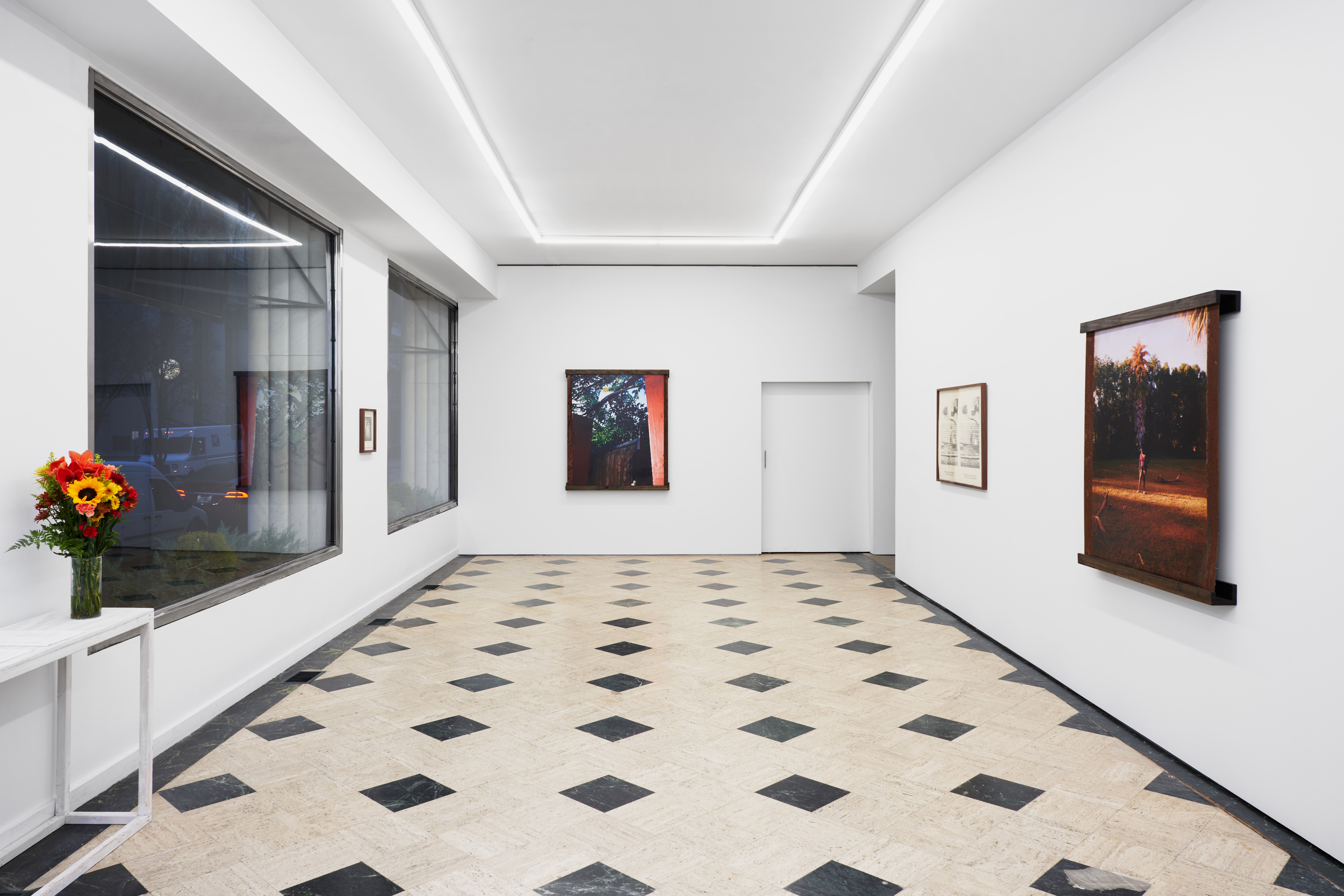 Brightly lit white wall gallery with large bay windows to the left, patterned black and white tile on the floor, and art lining the walls.