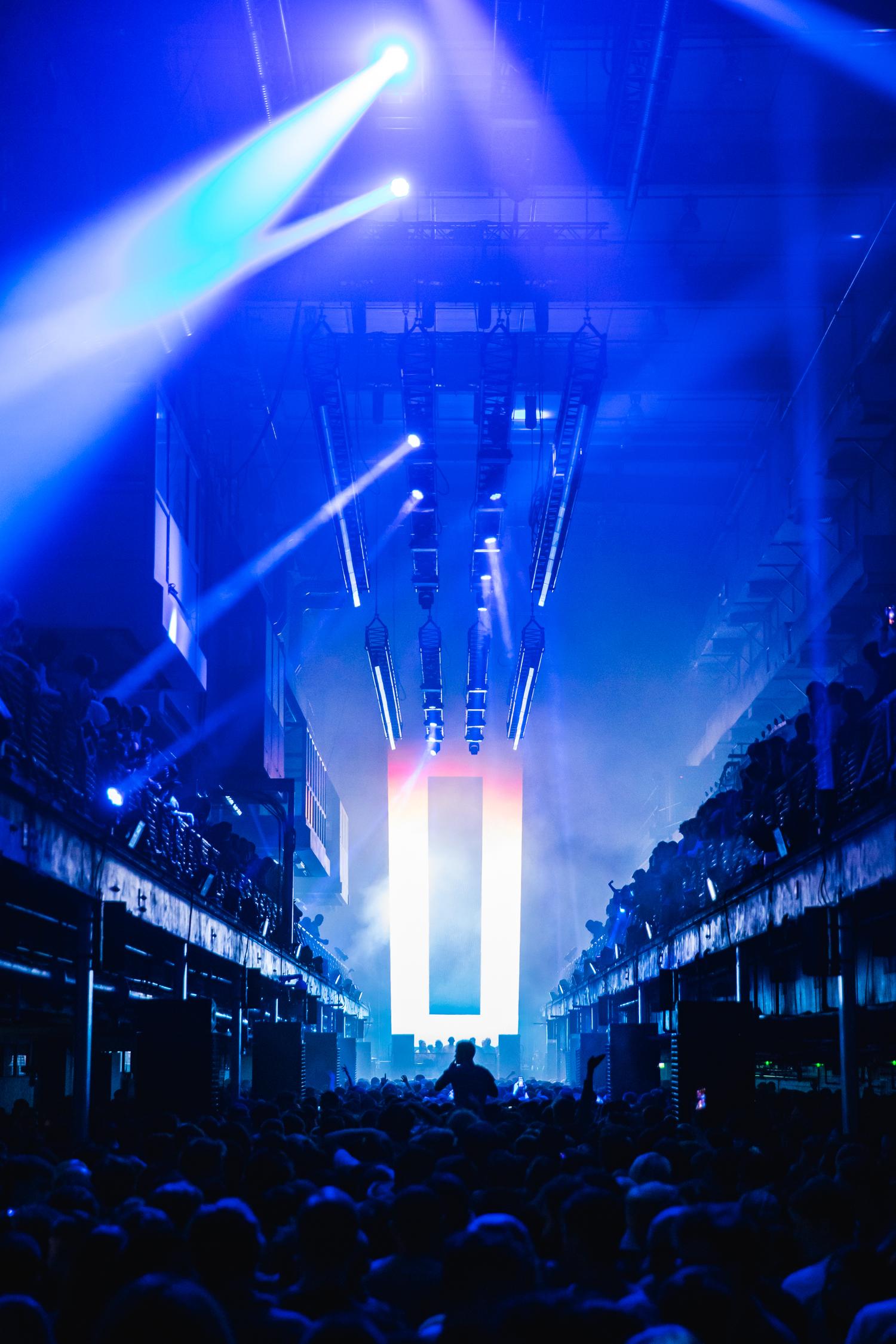 Blue stage lights illuminate Printworks London during AVA Club night with full crowd and DJ on stage.
