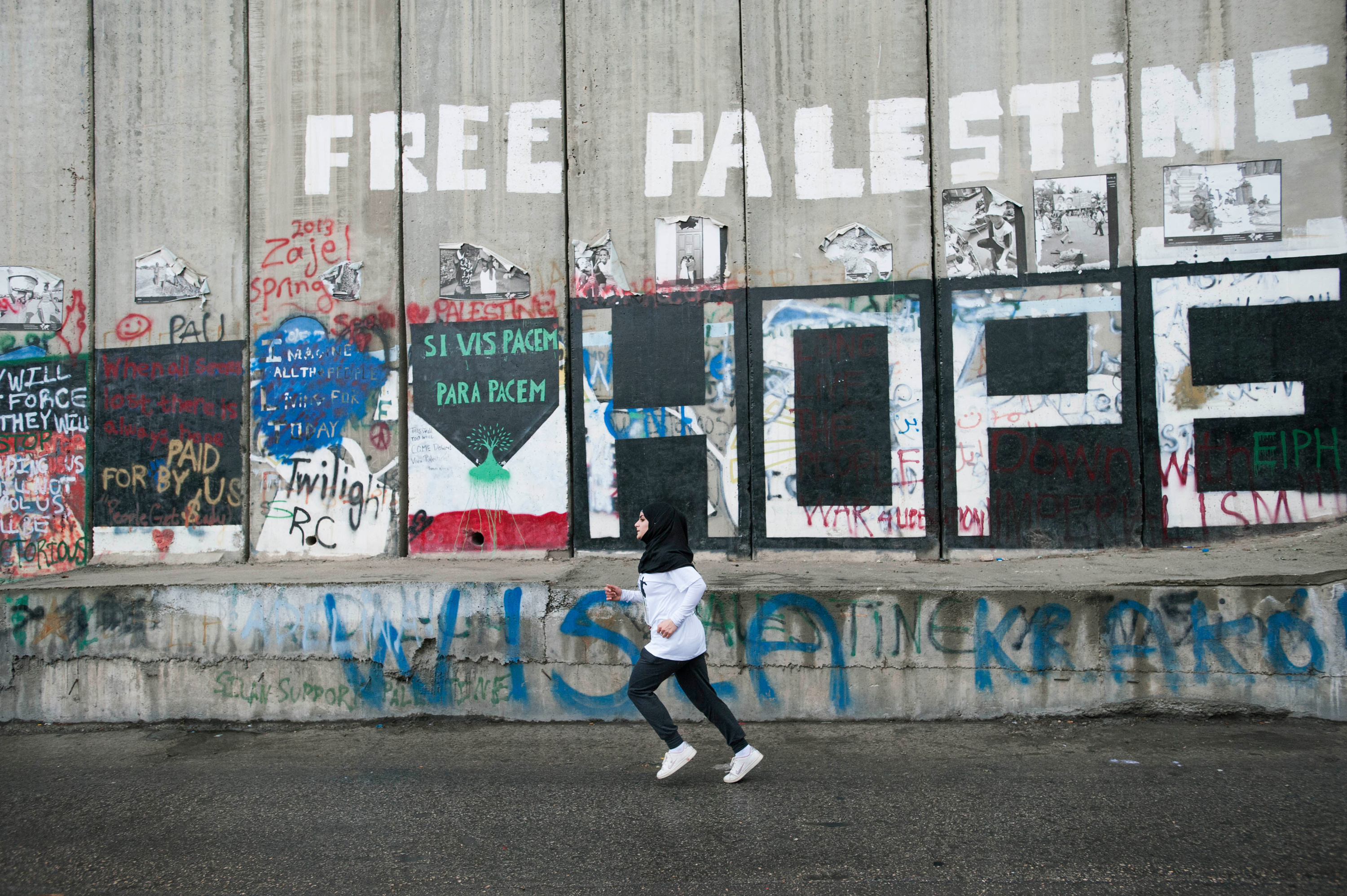 A single person seen from far away runs along a concrete wall graffitied with words like “Free Palestine” and “Hope” among many others.