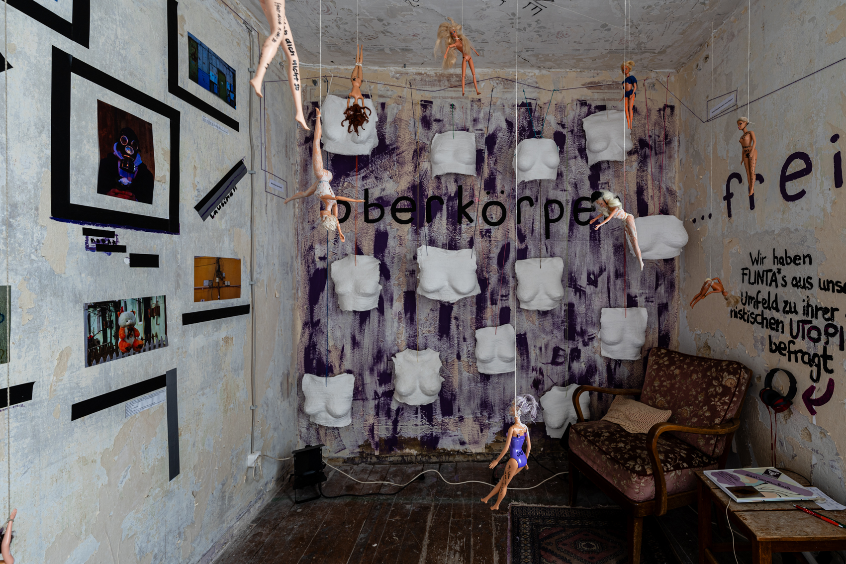 Bild 1 zu Patriarchat abnippeln: An der Wand gegen&uuml;ber h&auml;ngen Gipsabdr&uuml;cke von Oberk&ouml;rpern von Flintas. Die Wand ist teilweise mit breiten einzelnen lila Strichen bemalt. Darauf steht handgeschrieben "Oberk&ouml;rper". An der Wand rechts steht dann : "... frei". Darunter in der rechten Ecke ist ein alter Samtsessel. Neben ihm steht ein kleiner Tisch mit einem interaktiven Buch. An der Wand h&auml;ngen Kopfh&ouml;rer, auf welchen Utopien von Flintas zu h&ouml;ren sind.  Von der Decke h&auml;ngen beschriftete Barbiepuppen an Schn&uuml;ren. 
