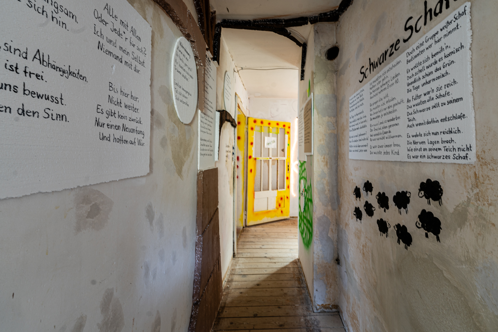 Bild 6: Zwischen schwarz und wei&szlig; Blick in den Gang zum Eingang der Wohnung: An den W&auml;nden stehen auf eckigen und runden Styroporfl&auml;chen Gedichte in schwarzer Handschrift. An der rechten Wand sind weitere kleine schwarze Schafe abgebildet. 
