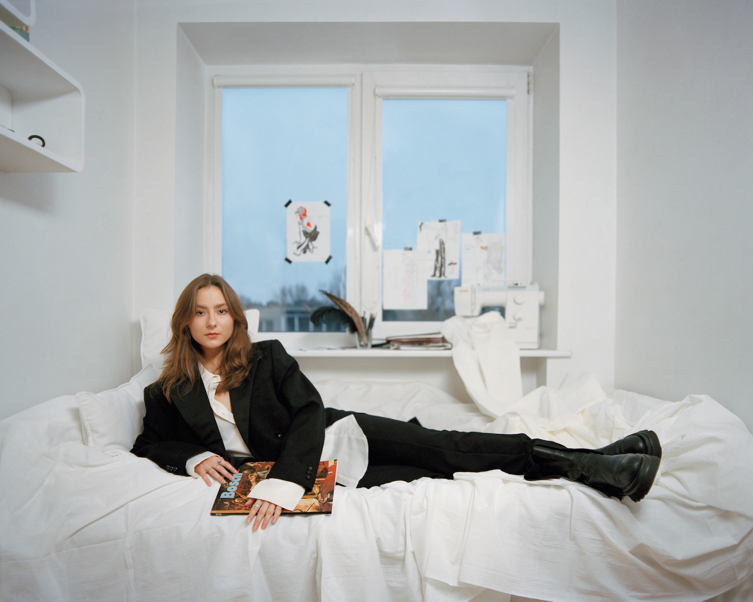 Emilija Poplavskytė, g. 1998 m. Emilija gimė ir užaugo Vilniuje. Jos tėtis buvo dailininkas Tadas Poplavskis, ilgą laiką kuręs ir gyvenęs Vokietijoje. Emilija nuo pat mažų dienų piešia ir domisi pasaulietinio meno istorija. Dabar mergina studijuoja Vilniaus dailės akademijoje mados kostiumo dizainą ir analizuoja ryšį tarp žmogaus psichologinių procesų ir išorinių pavidalų virsmo. Ji pasirinko fotografuotis savo baltame kambaryje, kuriame piešia ir kuria.