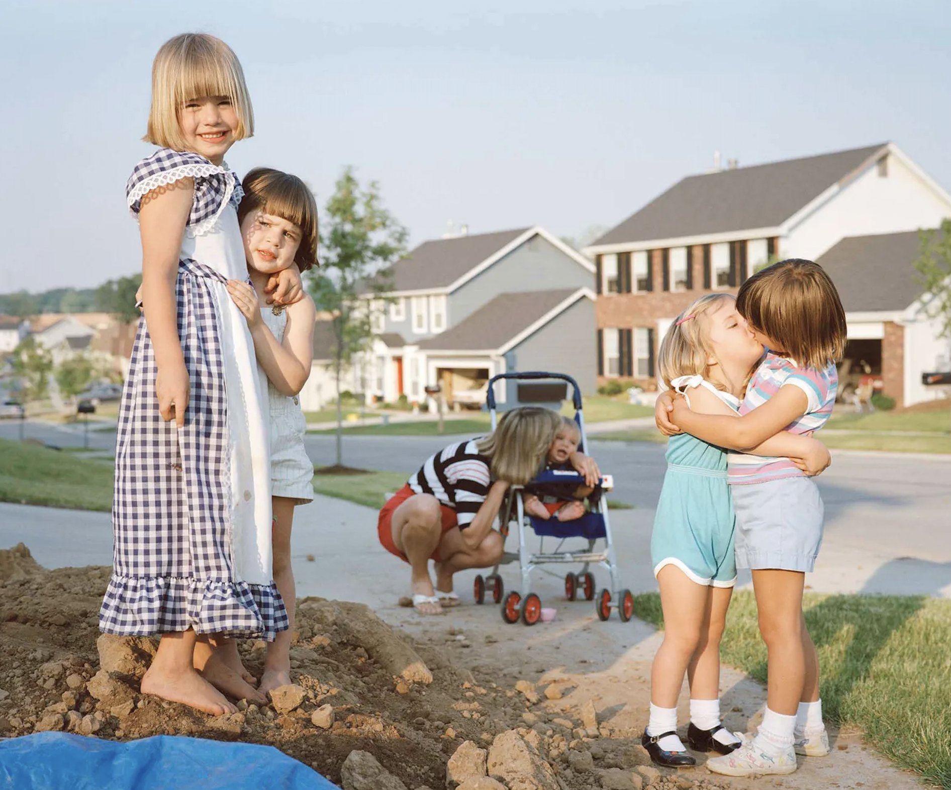Two children hugging and smiling into the camera, in the center background we see a mom kissing a child in a stroller, and to the left we see two little children kissing and embracing arms.